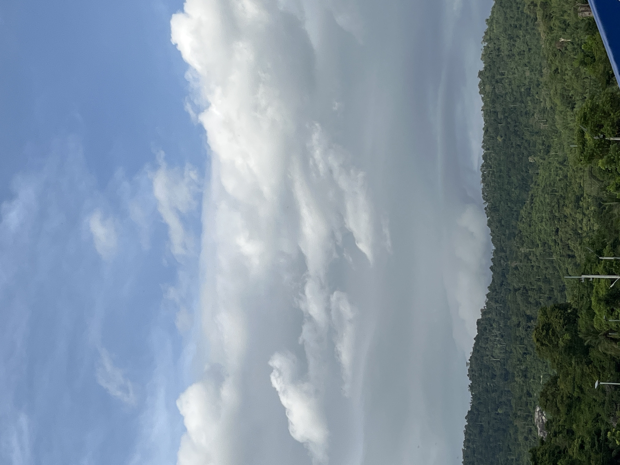 The image captures a scenic view in Chao Phraya Surasak, Thailand, featuring a combination of natural and man-made elements. Lush green foliage covers a steep, mountainous terrain. To the left of the mountains, the sky transitions from clear blue to dense clouds, suggesting potentially changing weather conditions. Electrical poles and wires are visible in the lower right corner, juxtaposed with the natural landscape.