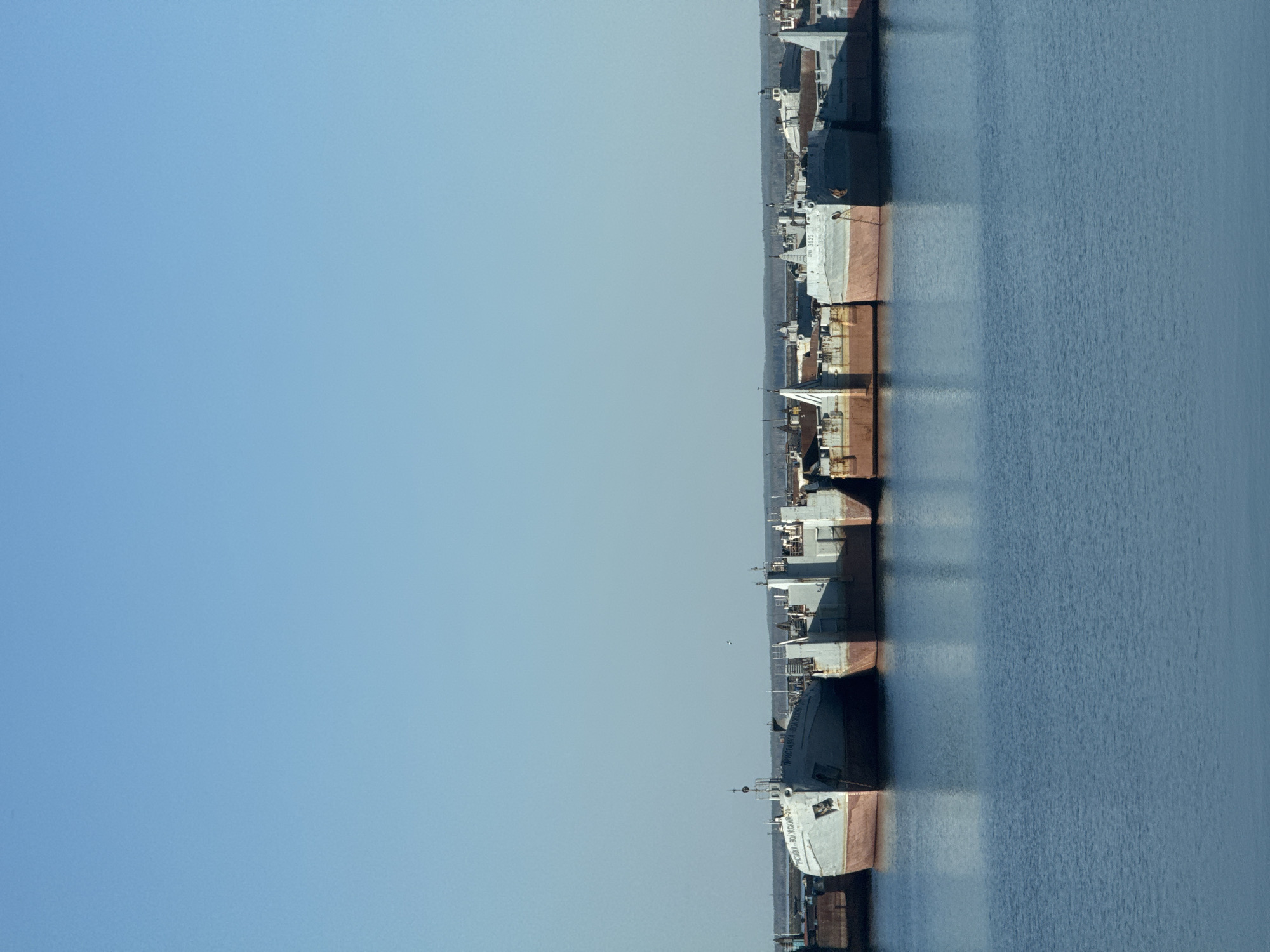 The image shows a series of ships docked along a river or harbor. The ships are lined up in a row, docked side-by-side. They appear to be cargo ships or large vessels, and each has a unique structure and design. The water of the river is visible beside the ships. A clear, blue sky fills the left side of the image. Given the location in Chaikovsky, Russia, it is plausible that the ships are docked on the Kama River or one of its tributaries. The image lacks any indication of people or activity at the time the photo was taken.