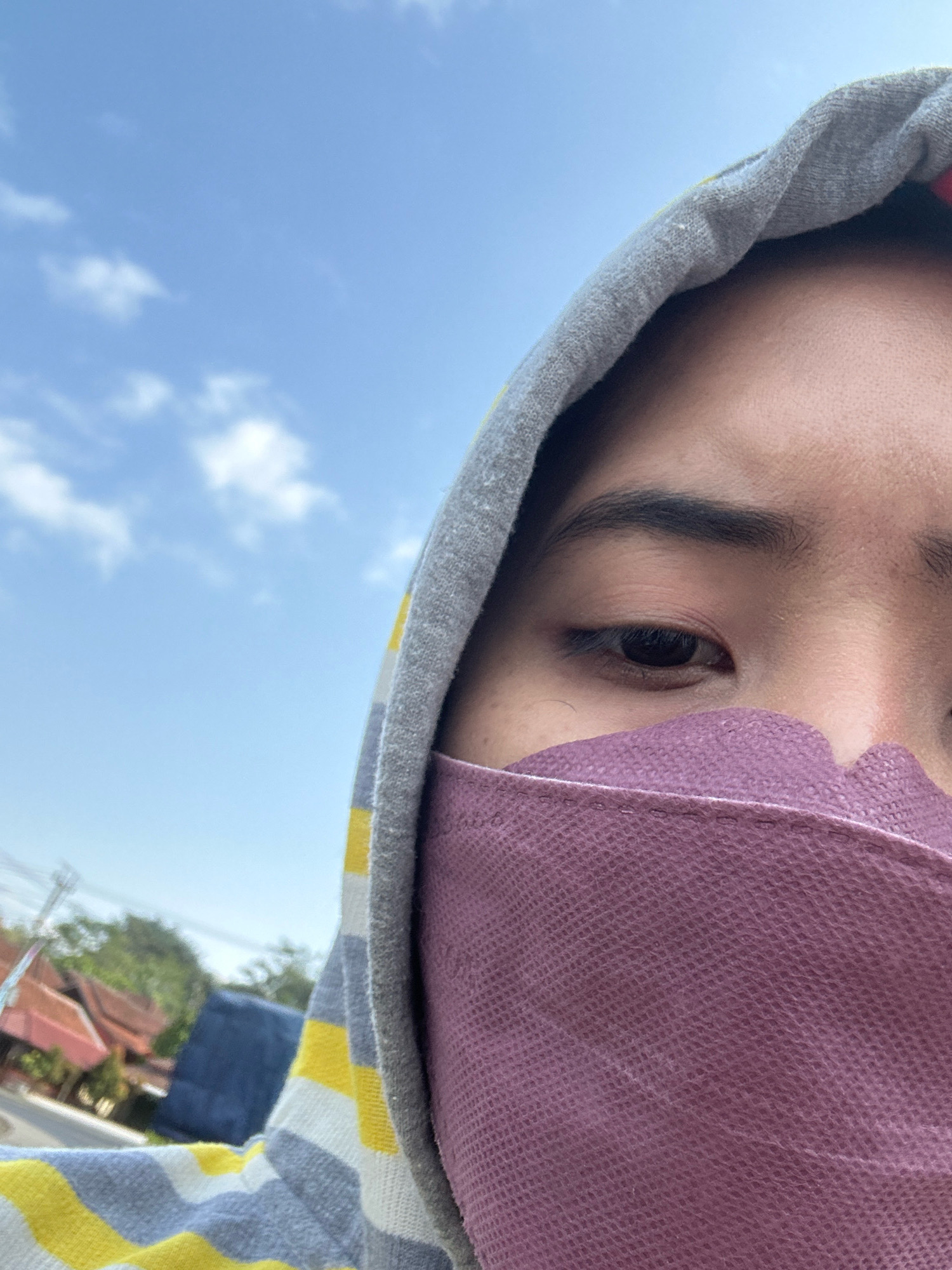 A close-up, low-angle shot shows the right side of a person's face, including their eye, eyebrow, forehead, and the upper part of their nose, which is covered by a purple mask. The person is wearing a grey hooded jacket with yellow and white stripes. The background is a clear blue sky with sparse white clouds. In the lower left corner, out of focus, are some buildings with terracotta roofs, trees, and a blue tarp, possibly indicating a residential area in a tropical climate. The scene appears to be outdoors during the daytime. The image does not provide specific information to pinpoint the location to Cihaurbeuti, Indonesia, but the architectural style and vegetation are consistent with tropical regions of Southeast Asia.