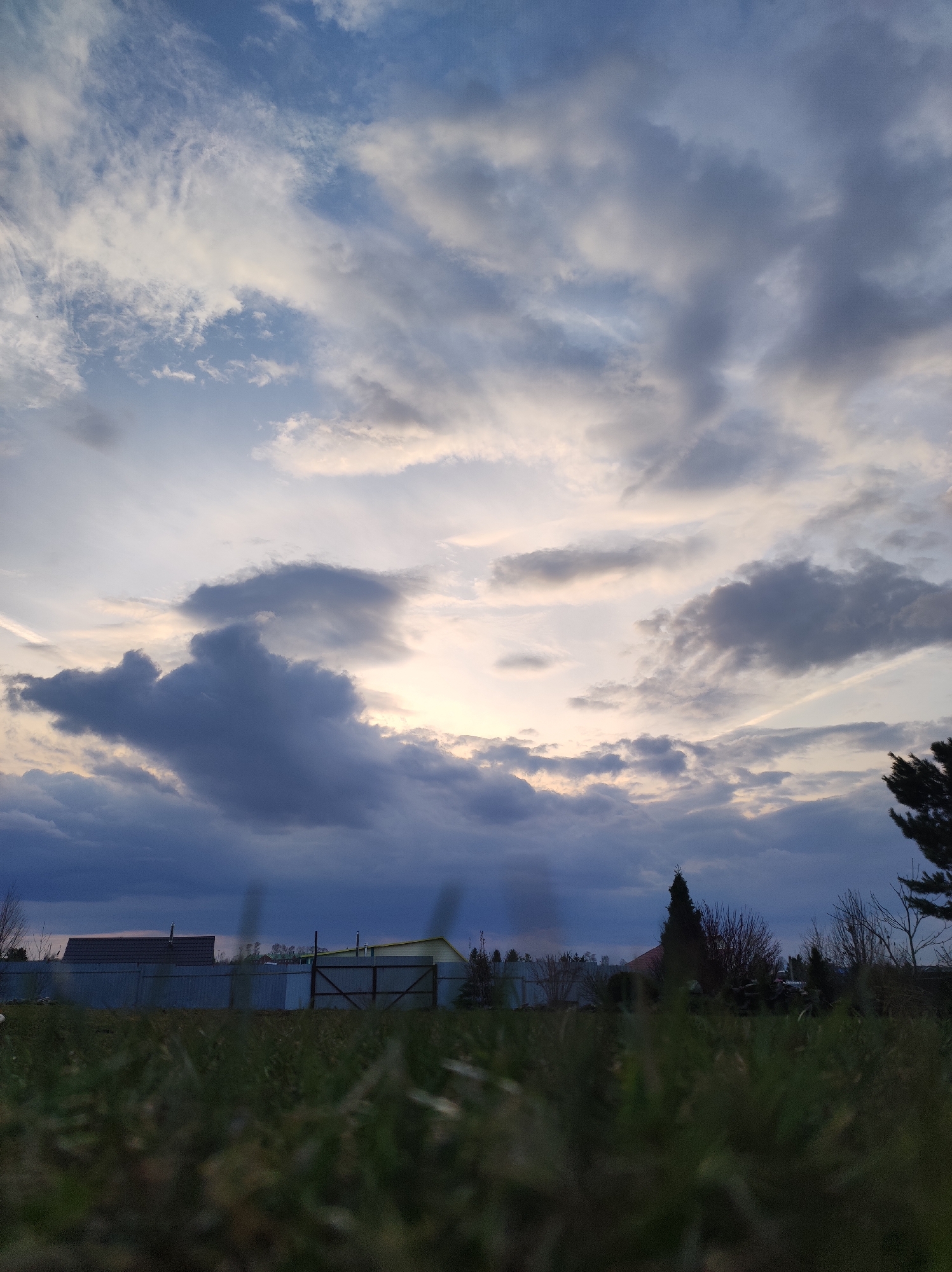 The image shows a view from ground level, likely within the urban landscape of Фоки, Russia. Dominating the background are houses obscured by a tall, grey fence. The foreground is dominated by grass, with the sky taking up the majority of the image. The sky features a complex array of clouds in various shades of grey and blue. Some light is visible behind the clouds. No people are visible in the image.