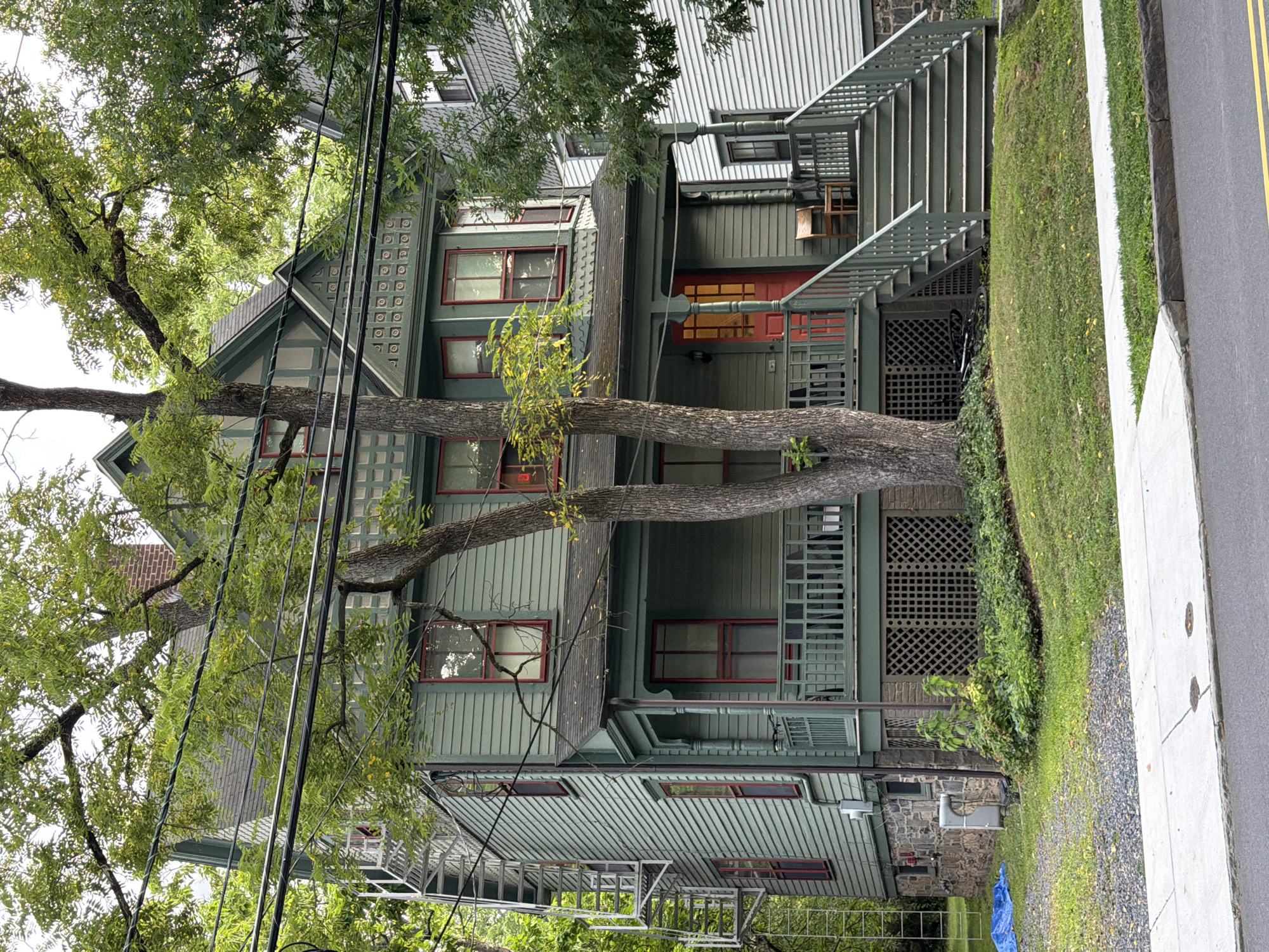 A two-and-a-half-story, green-sided house with red-trimmed windows and a covered porch is pictured on a cloudy day in Princeton, United States of America. A large tree with green foliage partially obscures the front of the house. A white metal fire escape is visible on the left side of the house. A set of gray wooden stairs leads up to the porch, which has a dark railing and a red front door. A wooden chair sits on the porch to the right of the door. The house is situated on a grassy lawn, with a sidewalk and a street in the foreground. Power lines are strung across the top of the image.