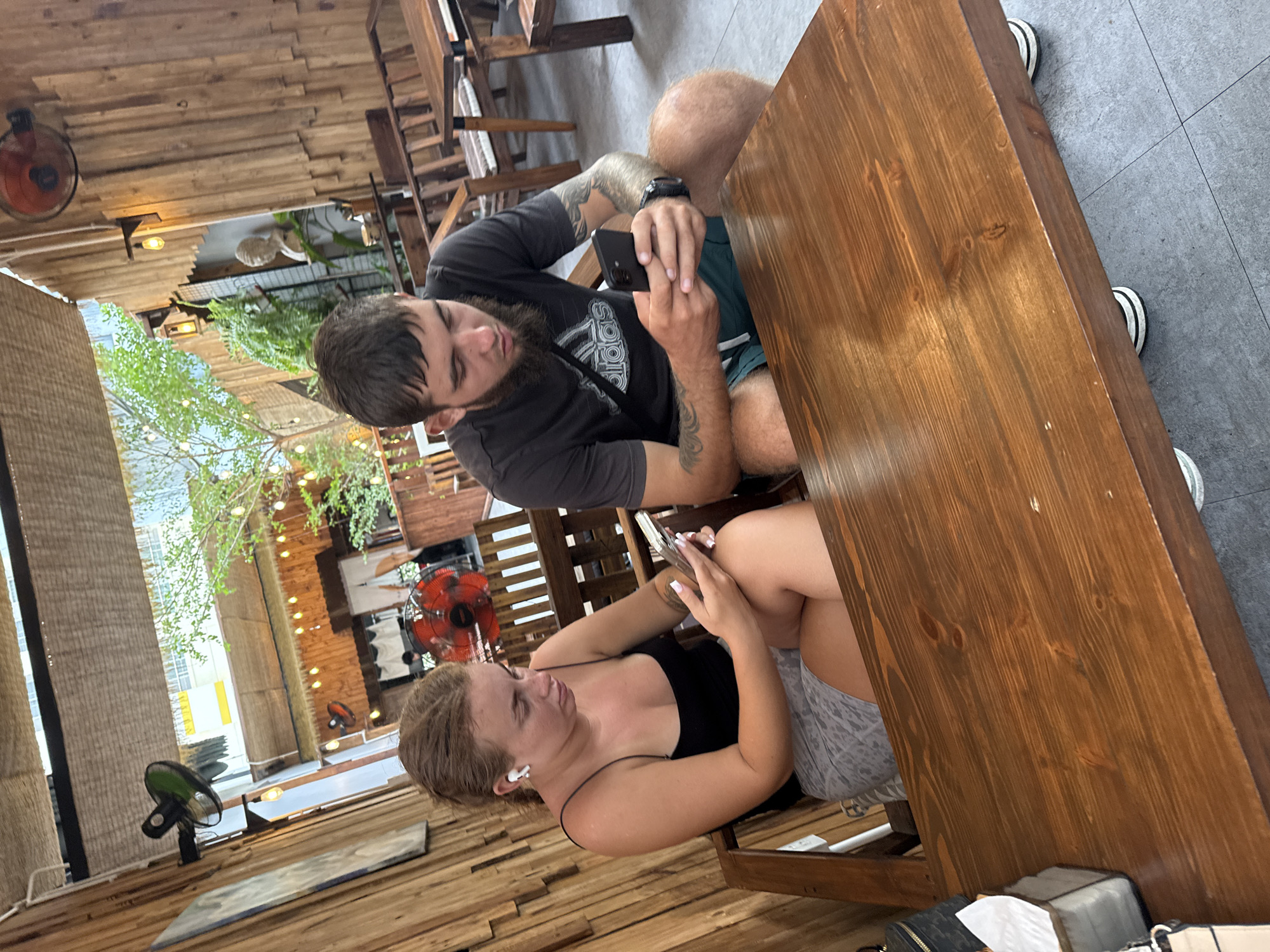 A man and a woman are seated at a wooden table in a cafe in Nha Trang, Vietnam. The woman, on the left, wears a black tank top and grey shorts, with white wireless earbuds in her ears. She is looking down at her mobile phone, holding it in her hands. The man, on the right, has a beard and is wearing a dark t-shirt with an Adidas logo and green shorts. He is also looking at his mobile phone, holding it in his right hand. Both individuals appear to be engaged with their devices. The cafe features wooden walls and furniture, including benches and tables. In the background, there are ceiling fans, string lights, and lush green plants, suggesting an outdoor or semi-outdoor seating area. A statue bust is visible on a shelf in the background. The floor is tiled.
