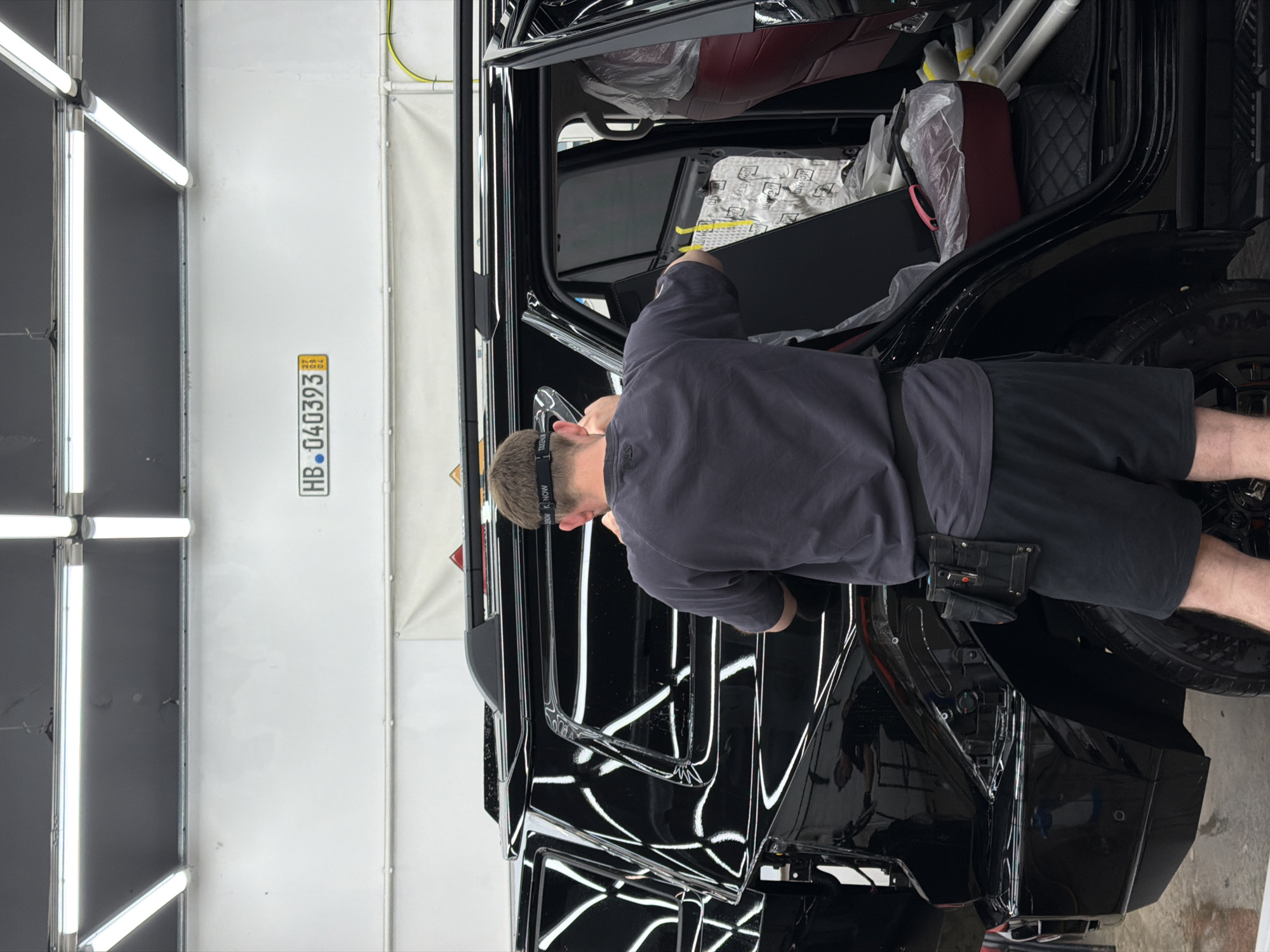 The image shows a man working on the interior of a black vehicle, possibly a luxury SUV, inside a well-lit garage or detailing shop. The man is wearing a gray shirt, black shorts, and a headband, and he appears to be focused on his work. The vehicle's interior features red seats and various parts covered in protective plastic. The environment is clean and organized, with fluorescent lights lining the ceiling. A license plate with the code "HB-040393" is visible on the wall. The code corresponds to the location of Krasnoyarsk, Russia.