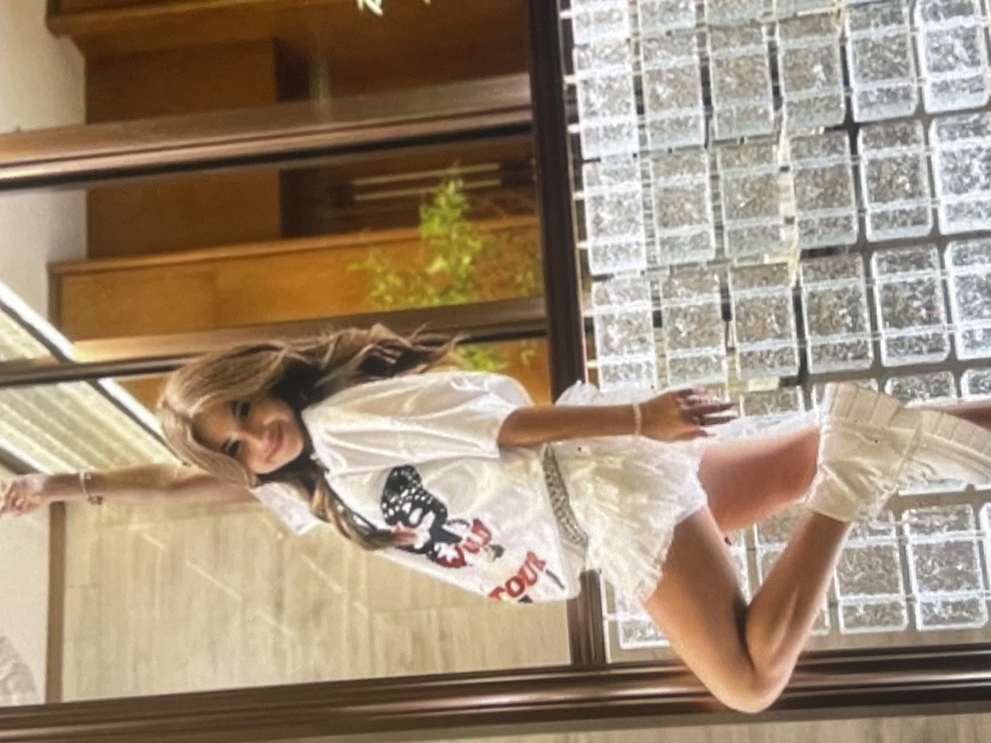A young woman is captured in a playful and dynamic pose, seemingly suspended horizontally against a large window or glass partition. She has long, light brown hair and a cheerful smile directed at the viewer. Her arms are outstretched, and her legs are bent at the knees, wearing white platform boots.
She is dressed in a casual yet stylish outfit, consisting of a white t-shirt featuring a graphic design of a black and white winged skull with a red element, and the partial text "...TOUR" clearly visible in red below it. She also wears a short, white, ruffled or lacy mini-skirt and a delicate bracelet on her left wrist.
The background shows an indoor setting with architectural elements. Through the clear glass of the partition, wooden structures resembling stairs or shelves are visible, along with some green foliage from a plant. To the right of the woman, there is a wall or partition constructed from textured, translucent glass blocks or tiles, creating a geometric pattern. A dark frame outlines the glass panel in the foreground.
The image appears to be taken during the daytime, with ample natural light. The woman's expression conveys happiness and confidence, contributing to a vibrant and energetic mood. The location context provided is Poltava, Ukraine.