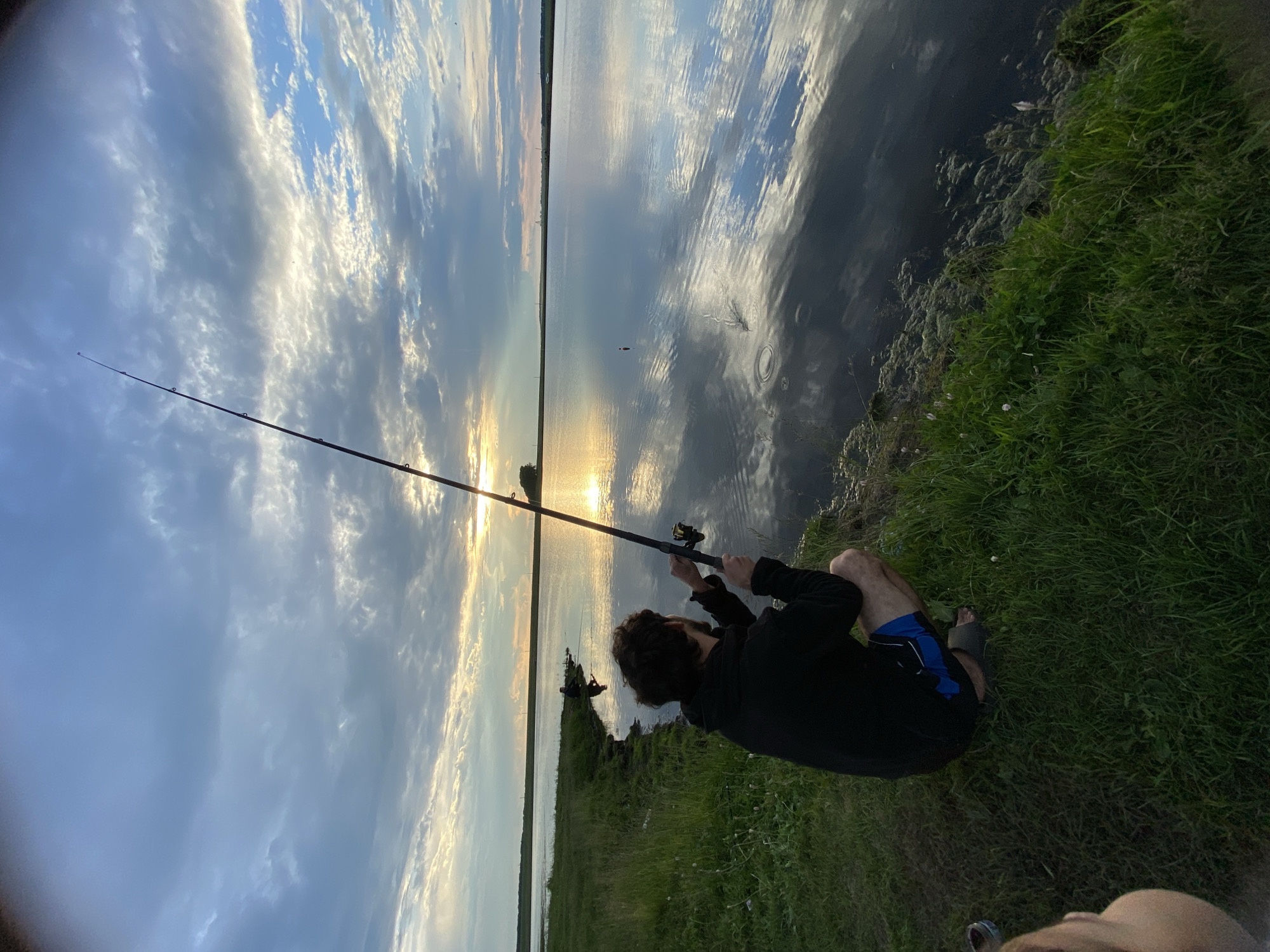 The image depicts a serene natural landscape with two individuals engaged in fishing. In the foreground, a man with dark, curly hair is crouched on the grassy bank of a tranquil body of water, holding a fishing rod. He is wearing a black hooded sweatshirt and blue shorts. His gaze is directed towards the water, where his fishing line is cast. Further in the background, another person is also fishing near the shoreline. The sky above is a canvas of dramatic clouds, with patches of blue sky and the warm glow of the setting sun breaking through, reflecting on the calm surface of the water. The environment suggests a rural or natural setting, possibly a lake or reservoir, located in the vicinity of Сыйрышбашево, Russia. The scene is peaceful and evokes a sense of leisure and connection with nature.