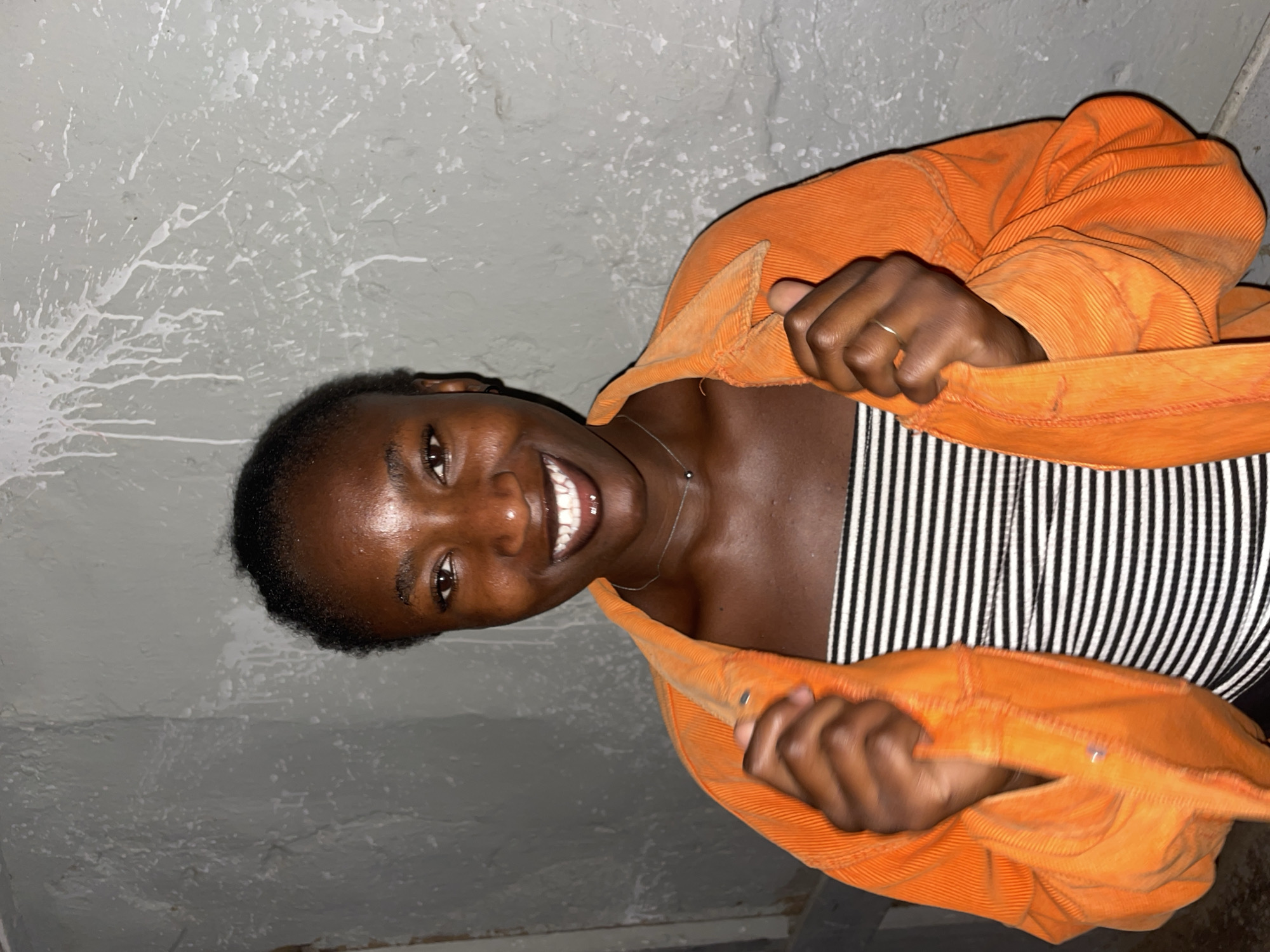 A smiling young woman is indoors, near a wall. The wall appears to have white marks or splatters on it. The woman has short, dark hair, dark skin, and is wearing a black and white striped top. She has a thin silver necklace and a ring on her finger. She is also wearing an open orange jacket or shirt made of corduroy-like material. The location is Jos, Nigeria.