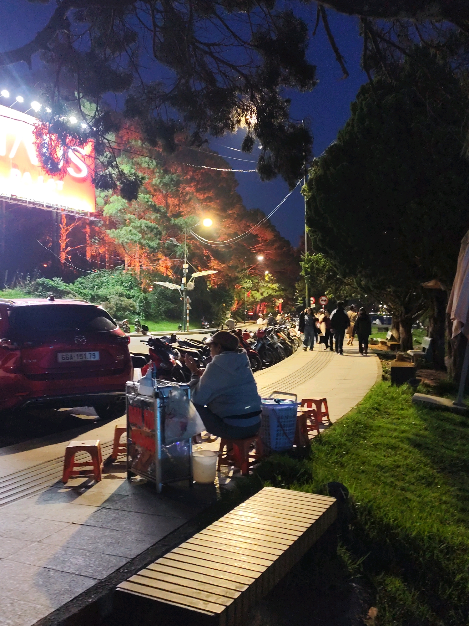 The image captures a scene at night in Dalat, Vietnam, likely on a street lined with trees and a sidewalk. In the foreground, a person is seated on a red stool next to a food cart. The cart appears to be metal with a glass display and various items inside. To the left, a red SUV with license plate 66A-151.79 is parked along the street. Further down the sidewalk, a group of people are walking away from the camera, silhouetted against the lights from a large billboard in the background. The billboard itself is partially obscured by tree branches and appears to be illuminated with bright orange and white lights, displaying what looks like text or graphics. Numerous motorcycles are parked along the side of the road. The environment is characterized by a dark sky, illuminated trees, streetlights, and the warm glow from the billboard, creating a lively evening atmosphere.