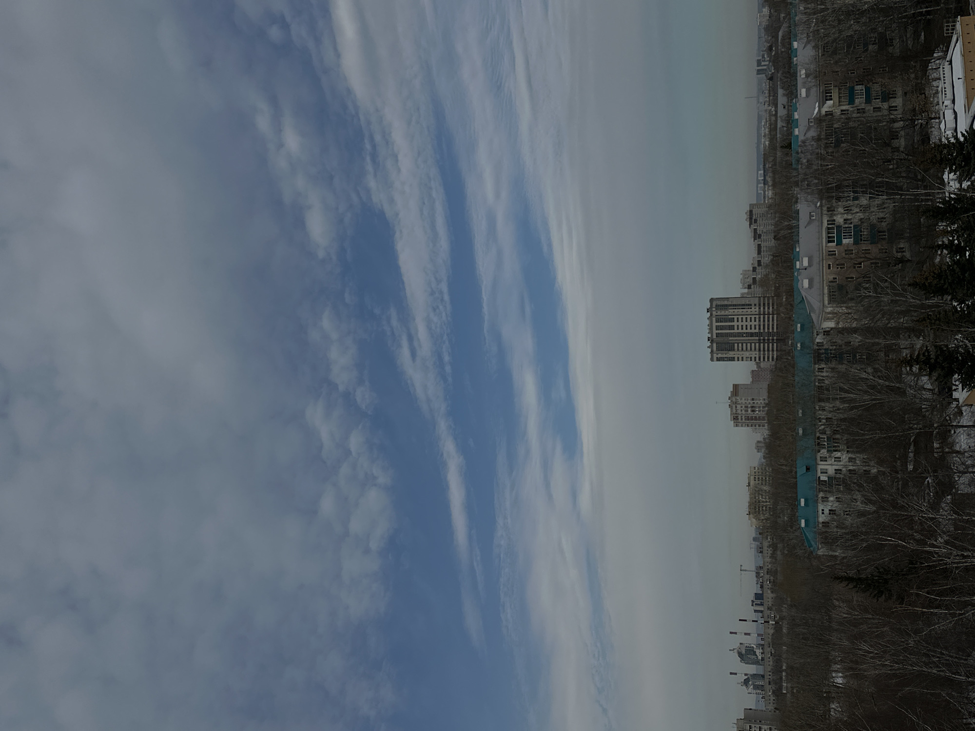 The image shows a cityscape of Kazan, Russia, under a sky with layered clouds. Several apartment buildings are visible, along with trees. Some of the buildings appear to be covered with snow. There are no people visible in the image.
