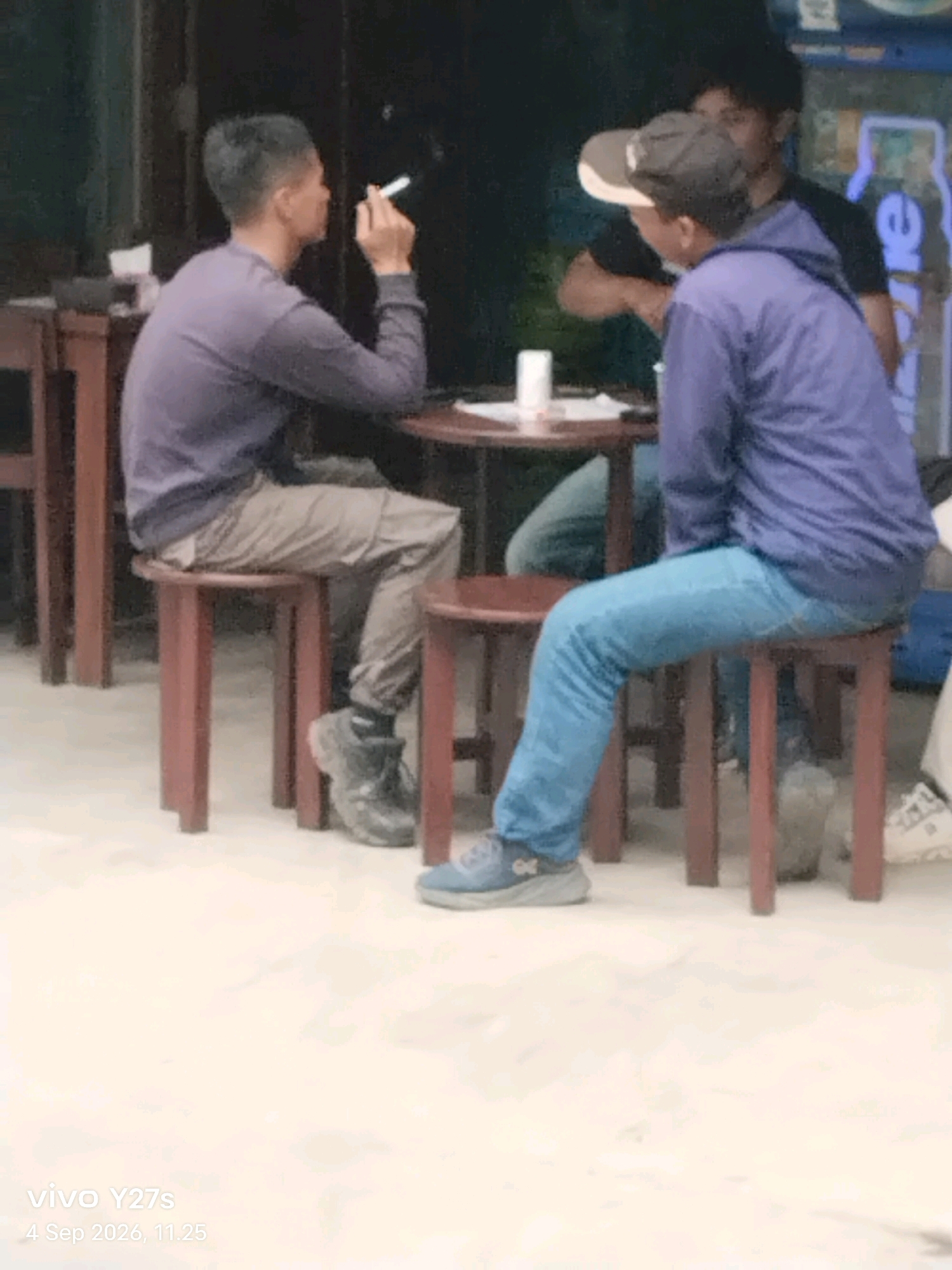 Three individuals are seated around a low table. The person on the left, facing right, wears a purple long-sleeved shirt and tan pants, smoking a cigarette. The person in the middle, facing left, wears a blue hooded jacket and blue jeans. The person on the right, facing left, wears a black t-shirt and a baseball cap. The setting appears to be an outdoor cafe or street stall with wooden stools and tables. The foreground shows a light-colored ground, possibly pavement, with a watermark indicating "vivo Y27s 4 Sep 2026, 11:25". The background is dark and indistinct, suggesting it might be nighttime or an indoor setting with low lighting.
