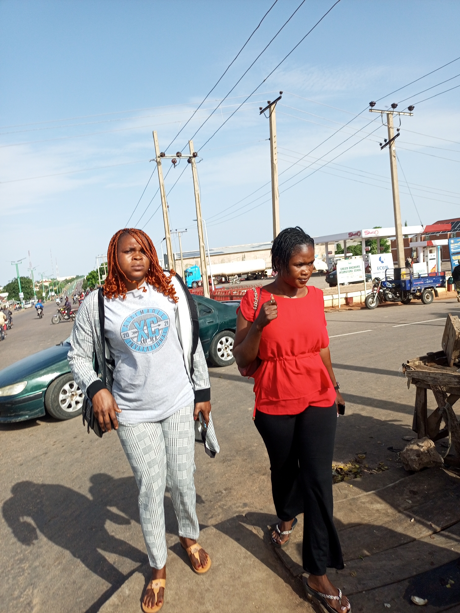 Two women are walking on a street in Tumfure, Nigeria. The woman on the left has on a grey t-shirt and pants and is wearing orange braids. The woman on the right is wearing a red shirt and black pants. In the background, there are cars, motorcycles, and utility poles with power lines. There is also a gas station with pumps and signs. The sky is clear and blue.