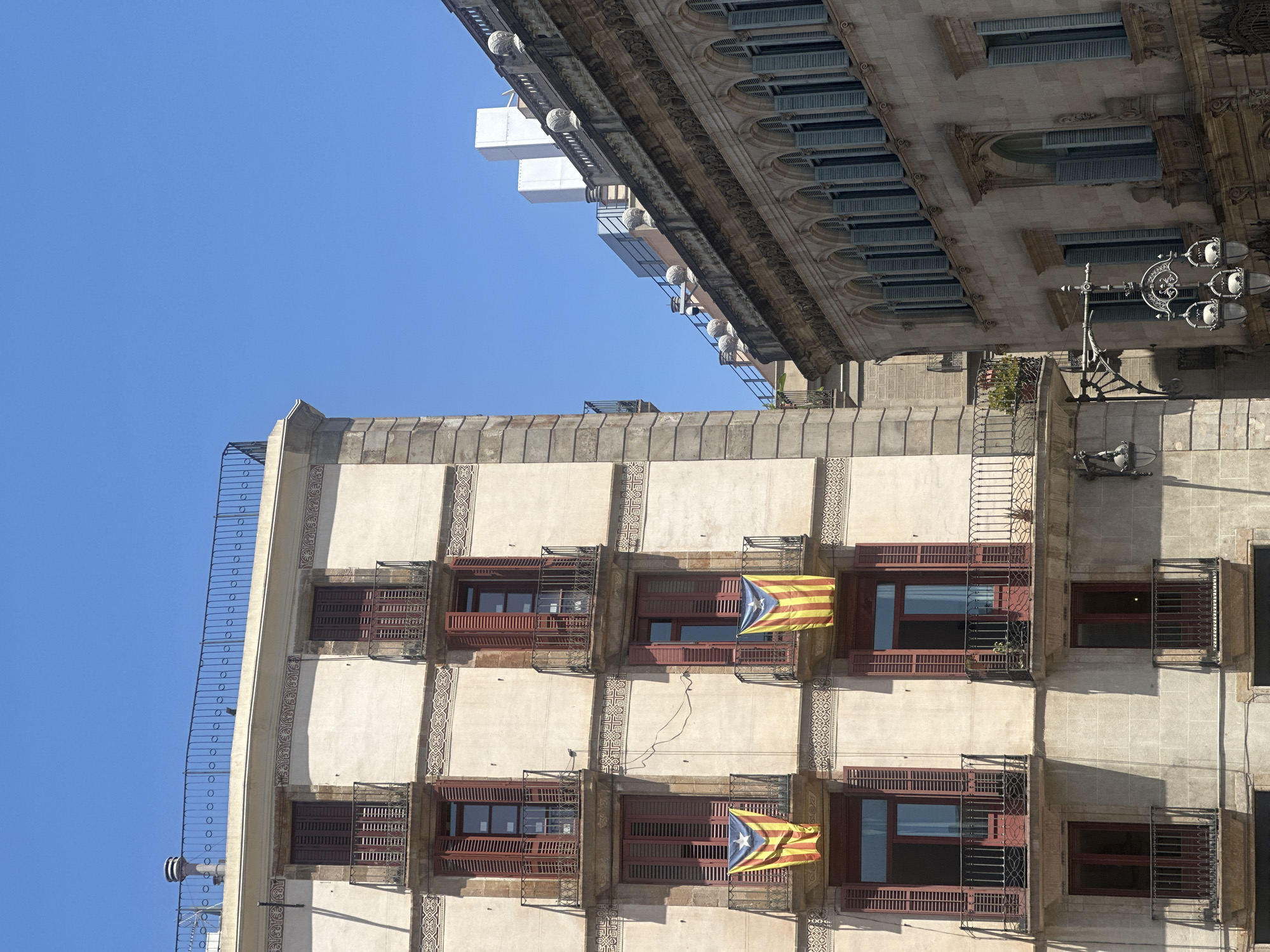 The image shows a close-up view of two buildings in Barcelona, Spain, under a clear blue sky. The building on the left is an older, multi-story structure with a light-colored facade. It features several balconies with dark-colored railings. Two Catalan flags, yellow with red stripes and a blue triangle with a white star, are hanging from balconies on the third floor of this building. The windows on this building have dark red shutters, some of which are open. Decorative trim adorns the building above the windows and between floors.
The building on the right is also ornate and appears to be made of stone. It has a series of arched windows with light blue shutters. The upper floors of this building are partially visible, revealing more architectural details and what appear to be rooftop structures.
In the foreground, an ornate street lamp with multiple light fixtures is attached to the side of the building on the right. The overall scene depicts a sunny day in an urban environment, likely a historic district of Barcelona. There are no people visible in the image.