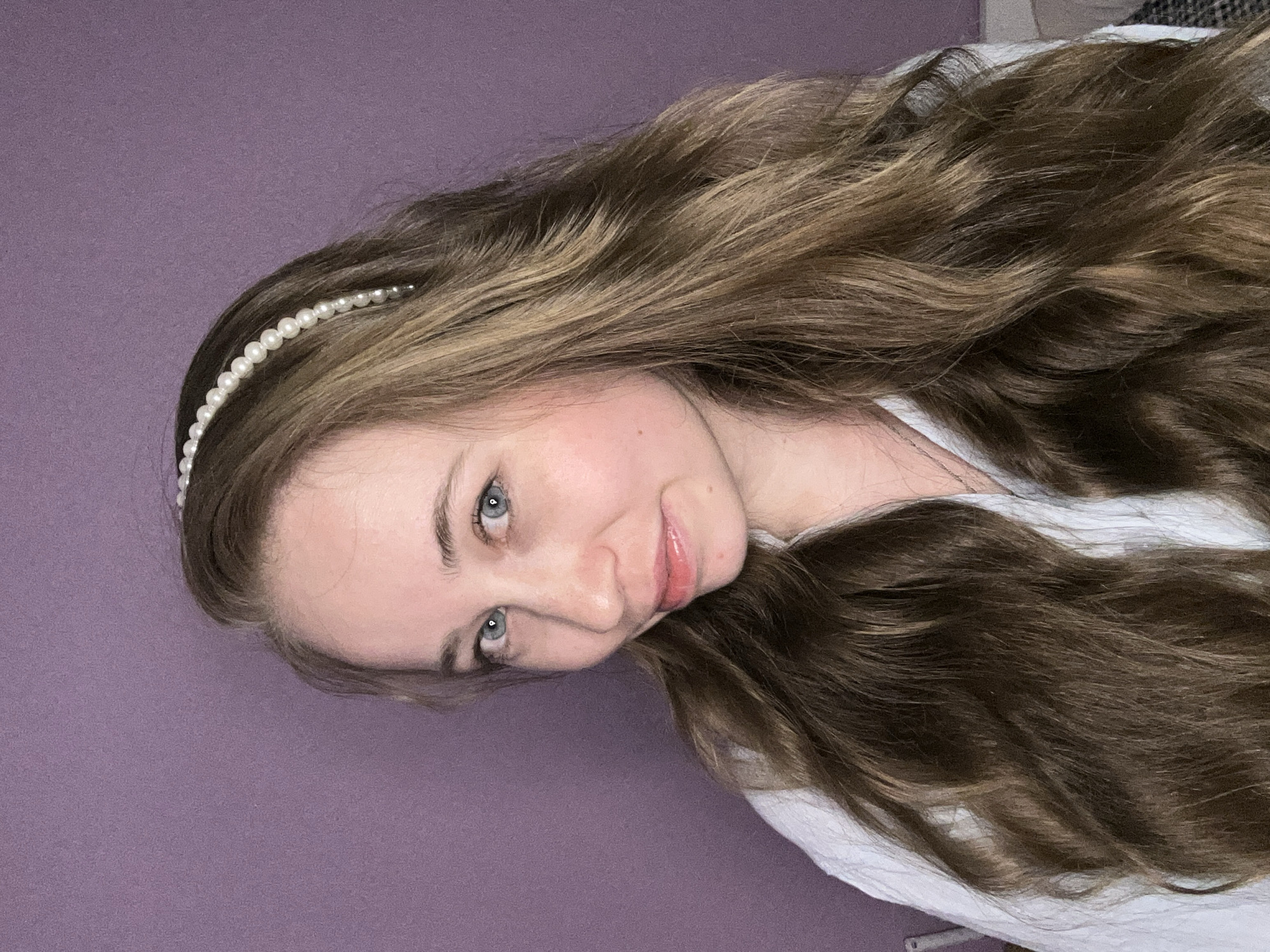 A young woman with long, wavy, light brown hair adorned with a pearl headband is posing against a solid, light purple background. She is wearing a white shirt. Her eyes are blue-grey, and she has fair skin. The scene is indoors. There is no evidence to suggest this image was taken in Saint Petersburg, Russia.