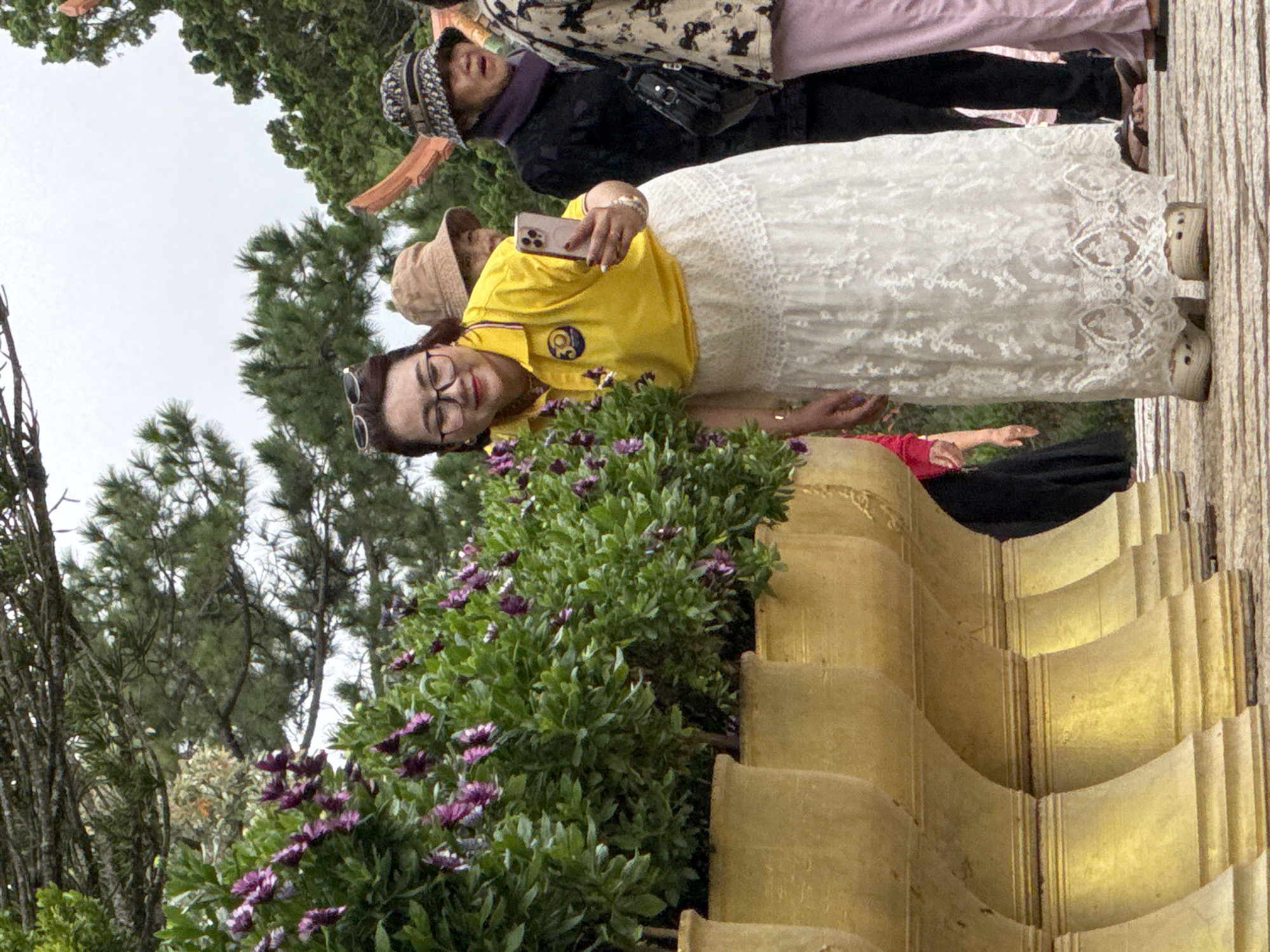 The image depicts a group of people outdoors, possibly at a tourist attraction in Da Lat, Vietnam. In the foreground, a woman in a bright yellow polo shirt and a long, white lace skirt is holding up a smartphone, seemingly taking a selfie or recording a video. She is wearing glasses and has a warm smile. To her right, two other individuals are partially visible: one wearing a beige bucket hat and another with a patterned hat and a dark scarf. Behind them, a lush display of green foliage with small purple flowers is prominent, contained within large, golden planters arranged in a row. The background suggests a natural setting with trees and a cloudy sky. The overall impression is of a pleasant, casual outing at a scenic location.