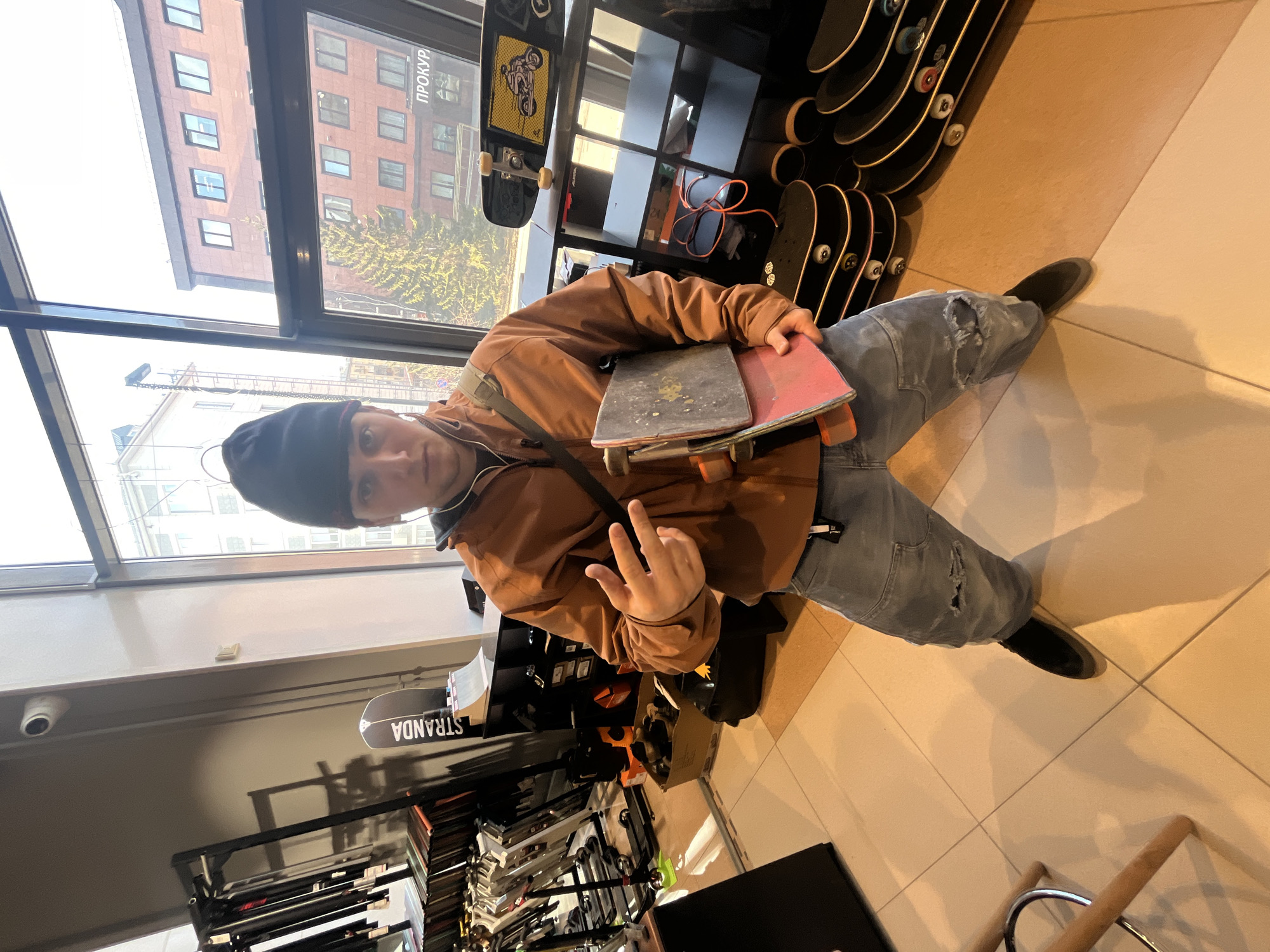 The image shows a young individual posing inside what appears to be a skate shop, looking directly at the camera. The person, possibly male, is wearing a dark beanie, a brown zip-up jacket, light blue ripped jeans, and dark shoes. They are holding two skateboards, one grey and one red, in their left arm, and making a peace sign with their right hand. A black shoulder bag strap is visible across their chest.
The setting is an indoor retail space with a light-tiled floor. Numerous skateboards are displayed on racks and shelves, some hanging vertically, others stacked horizontally. Other skate-related accessories and equipment are also visible on shelves in the background. A security camera is mounted on the ceiling.
Large windows dominate one side of the shop, offering a view of an urban environment. Through the windows, multi-story brick buildings and trees with sparse foliage are visible, indicating an outdoor setting, likely during autumn or early spring, illuminated by natural daylight. On one of the buildings across the street, the Russian word "ПРОКУРОР" (PROKUROR), meaning "Prosecutor," is prominently displayed. A skateboard displayed inside the shop also shows the word "STRANDA" written upside down. The activity captured is a casual pose within the store.