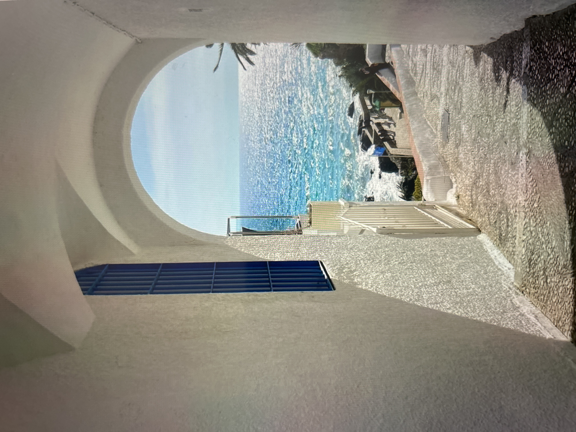 The image displays a picturesque coastal scene viewed through a striking white architectural passage. The perspective is from within a curved, white archway or tunnel, looking out towards a sparkling blue ocean under a clear sky.
In the immediate foreground, a smooth, off-white wall forms the left side of the passage, featuring a horizontal blue louvered window or vent. The archway frames the central view. On the right, a textured path, seemingly made of light-colored gravel or small stones, slopes downwards. A white metal gate is partially visible on the right, marking an entrance or boundary.
Beyond the archway, the main subject is a vibrant expanse of clear blue and turquoise sea, reflecting bright sunlight that creates numerous shimmering highlights on the water's surface. Gentle waves can be seen breaking against a rocky coastline on the right side of the ocean view. Green foliage and palm tree fronds are visible on the distant shore, and parts of structures or terraces are nestled along the rocks. Another palm tree frond extends from the upper left side of the arch's opening.
The scene is bathed in bright daylight, suggesting late morning or early afternoon, with clear weather conditions. Strong sunlight casts distinct shadows, enhancing the textures of the white walls and the gravel path. The architecture, with its white stucco and clean lines, combined with the vivid blue sea and sky, evokes a serene Mediterranean or tropical ambiance. There is no discernible activity or any visible text.