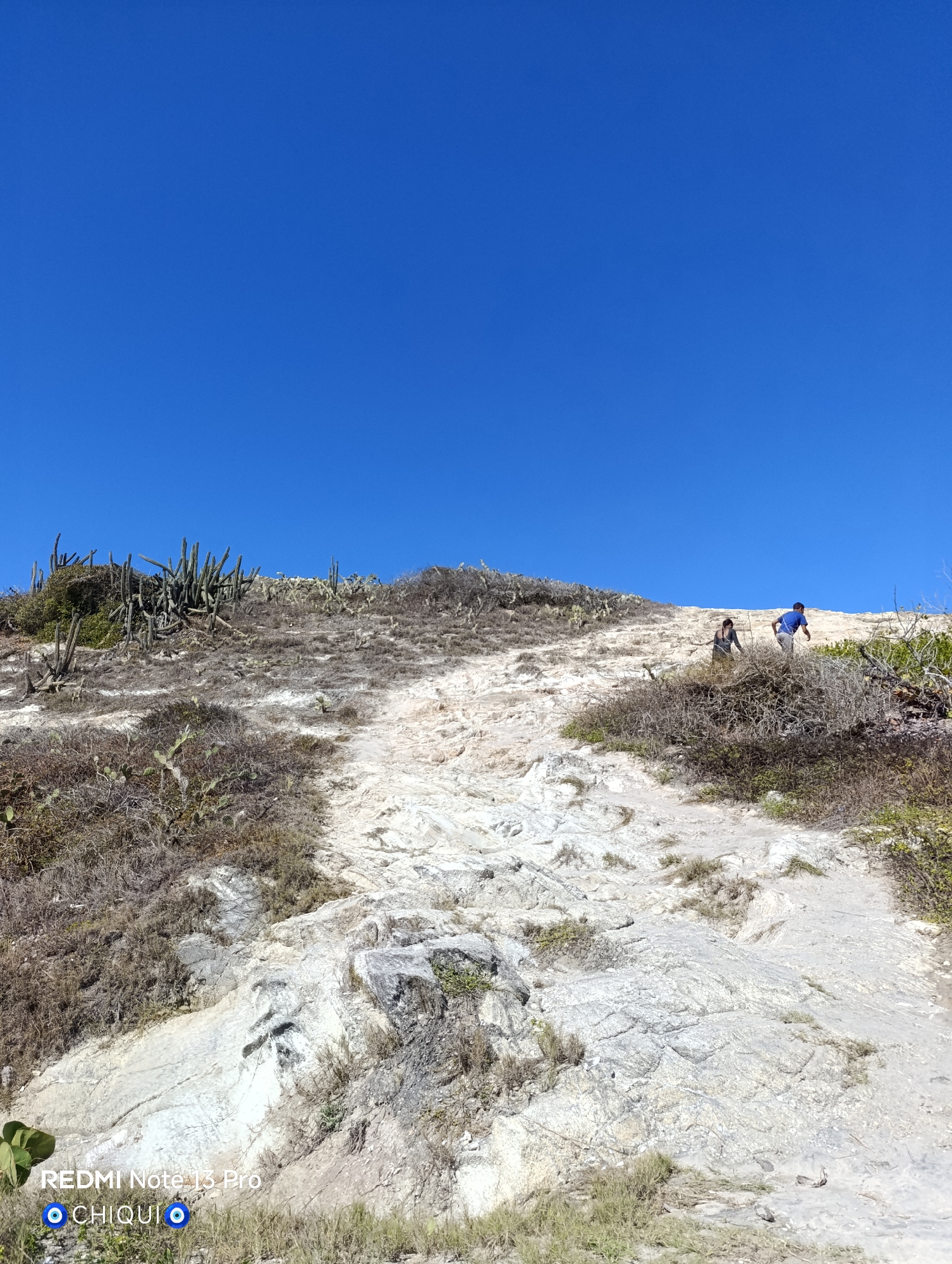 The image, taken in Playa Parguito, Venezuela, showcases a rocky, arid hillside under a clear blue sky. Two people are ascending the slope. One person is wearing dark clothing, while the other is in a blue shirt. The hillside is predominantly light-colored rock with sparse vegetation, including cacti. The foreground shows the lower part of the rocky path. A watermark reading "REDMI Note 13 Pro © CHIQUI ©" is visible in the bottom left corner of the image.