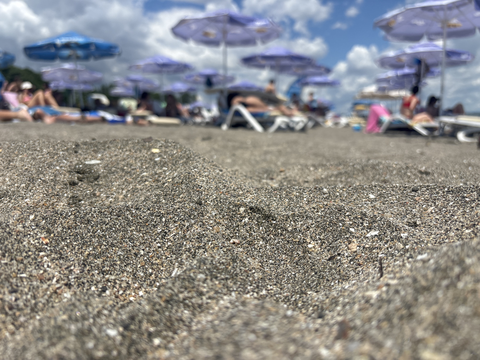 The image shows a close-up view of sand on a beach, with lounge chairs and umbrellas in the blurred background. The sand is dark gray and composed of small pebbles and grains. Several beach umbrellas, predominantly purple and blue, are visible, along with some lounge chairs and the silhouettes of people relaxing. The sky above is partly cloudy. The scene depicts a typical beach environment on a sunny day.