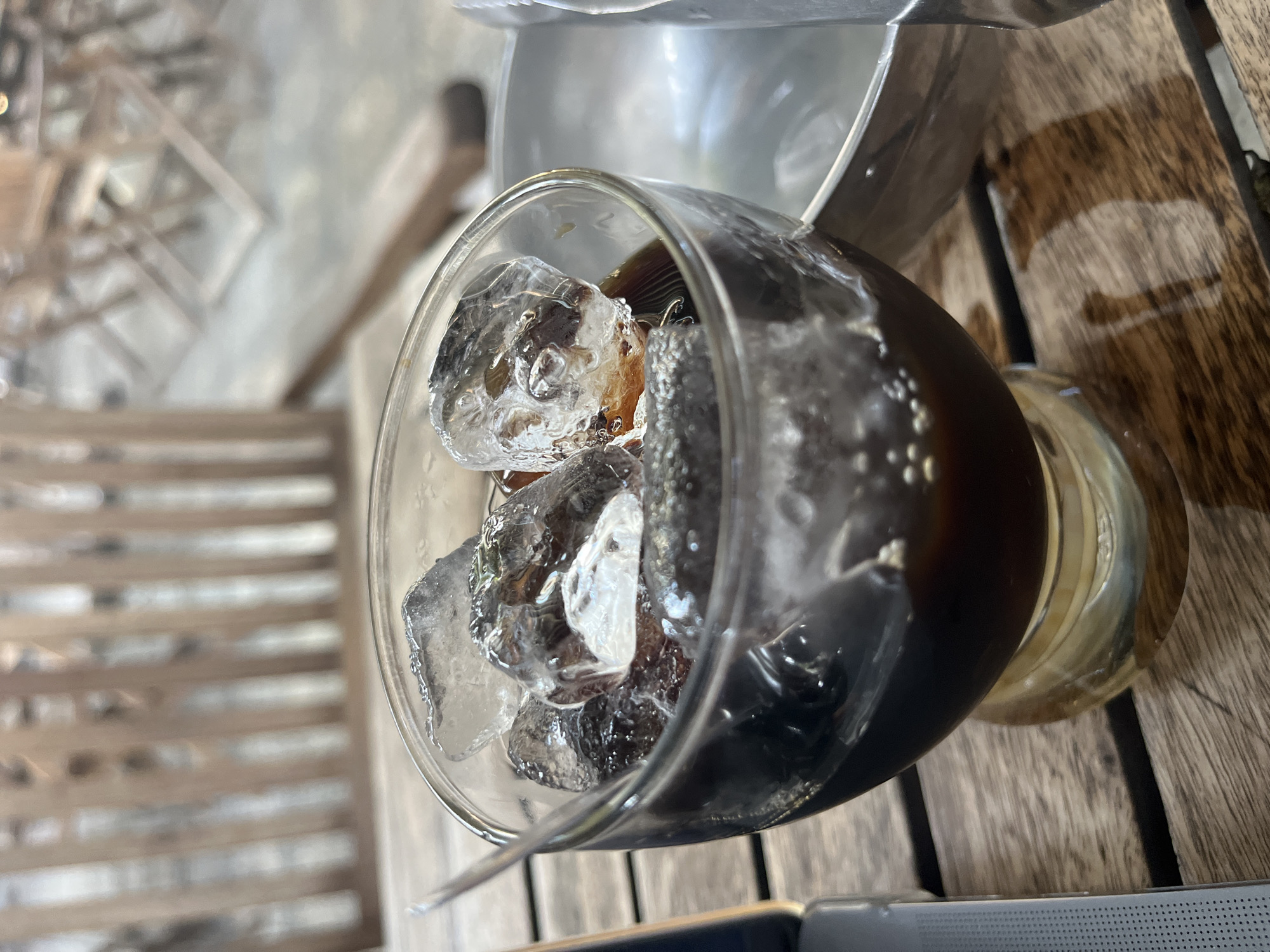 A glass filled with ice and a dark liquid, likely iced coffee, sits on a wooden surface. The glass is partially obscured by a silver container above it. In the background, blurred slats suggest a bench or other seating. The precise location within Nha Trang Ward, Vietnam is not determinable from the image, there are no people visible.