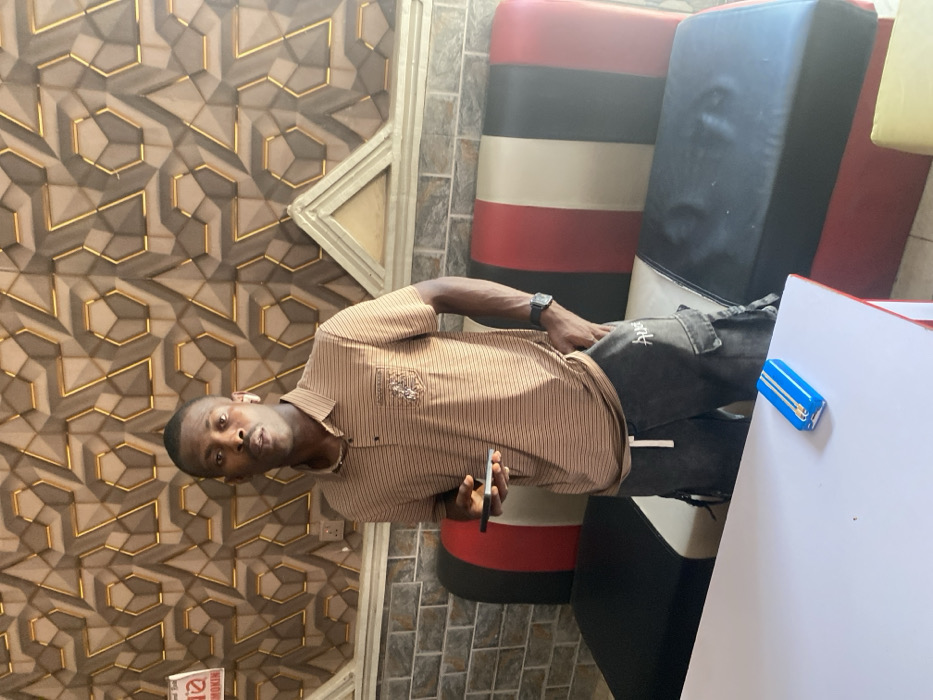 An individual, an adult male with short dark hair, a mustache, and a goatee, stands upright and faces the camera in an indoor setting in Zaki Biyem, Nigeria. He wears a light brown horizontally striped polo shirt with an emblem on the left chest and dark grey cargo shorts with a white drawstring and "Aug" text on the left thigh. A dark digital wristwatch is on his left wrist, and a white earbud is in his right ear. He holds a dark-colored smartphone in his left hand, screen up, while his right hand rests in his shorts pocket.
The background wall behind him features a textured, three-dimensional geometric pattern in brown/beige with gold outlines, positioned above a grey brick-tiled section. A decorative light-colored arch structure is present on the wall, along with a white electrical outlet and a partial red and white sign displaying "NIMOKIN" text in the upper left. To the man's right, a long bench-style seat is visible, upholstered in vertical sections of black, white, and red. In the foreground, a white rectangular table with red trim holds a small, rectangular blue object featuring a white zipper design on its top surface.