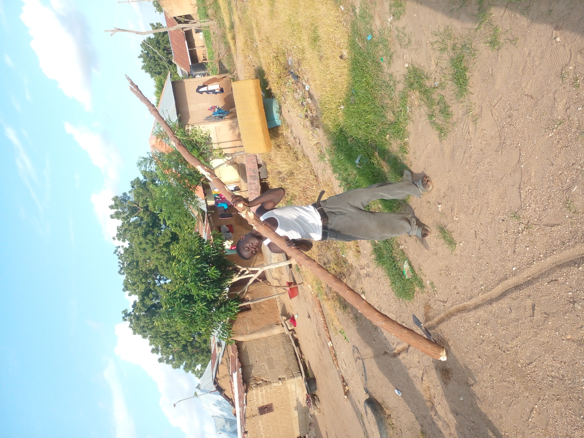 In a rural setting, under a clear blue sky with scattered clouds, a young Black man stands outdoors, holding a thick, long log at an angle. He is wearing a white tank top and loose-fitting grey pants. His body is tilted as he grapples with the weight of the log. The ground is a mix of dirt and patches of dry grass, with a visible tire in the background to the left. Scattered around the scene are several houses with corrugated metal roofs and mud brick walls. A tree with lush green foliage stands behind the man, and laundry is visible hanging on a line attached to one of the houses. The overall impression is of a scene of manual labor in a village environment.