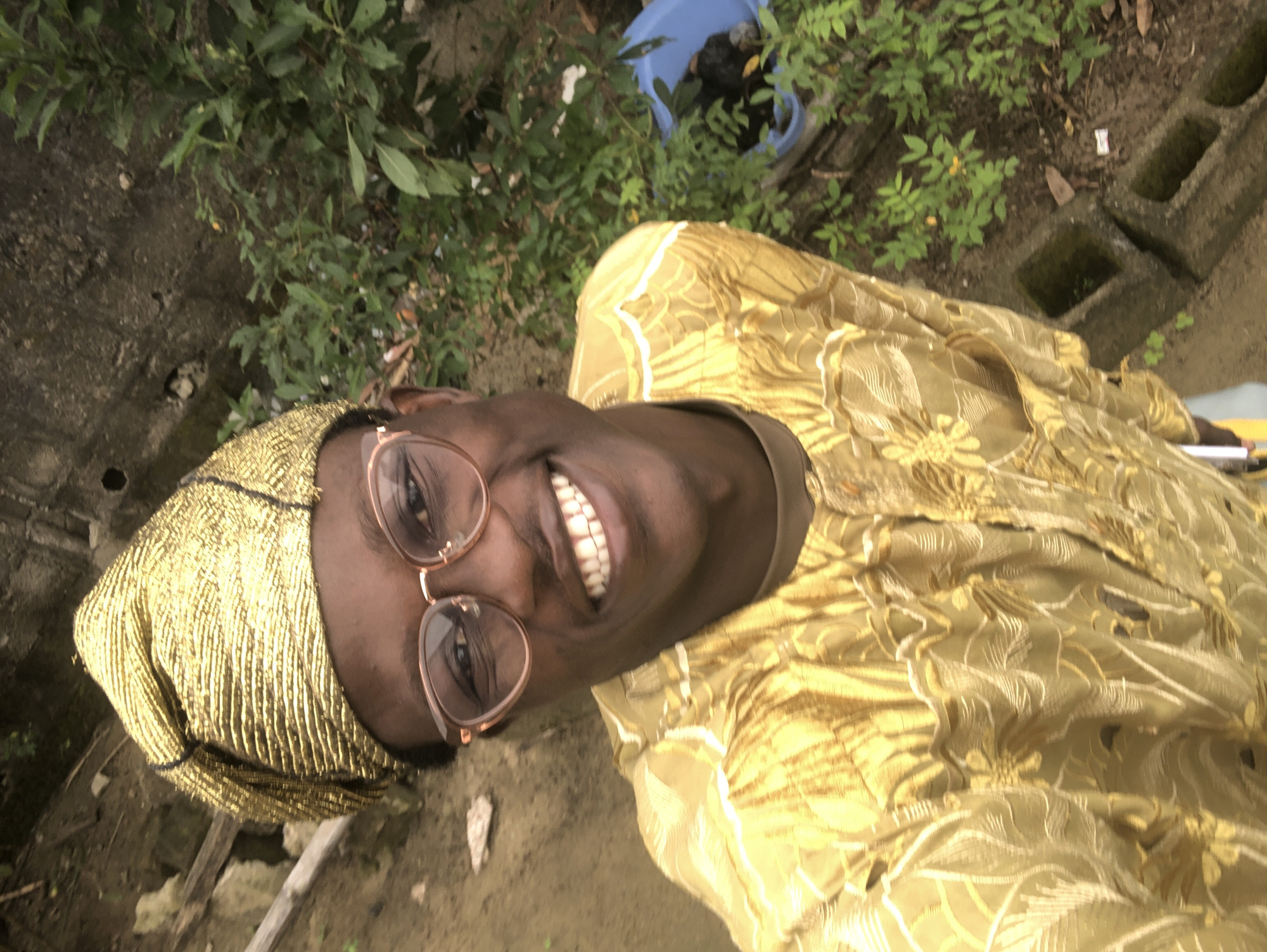 A person, wearing a gold headwrap and a gold, patterned garment, is captured in a selfie. The person is smiling broadly, showing their teeth, and wearing oval-shaped glasses with rose-gold rims. The background is outdoors, with lush green foliage and some dirt. In the background, a blue container and some concrete structures are visible, suggesting a possibly unkempt or rural setting. The lighting suggests it might be daytime, and the overall atmosphere seems relaxed and cheerful. The image appears to be taken in Ado, Nigeria. There is no visible text in the image.