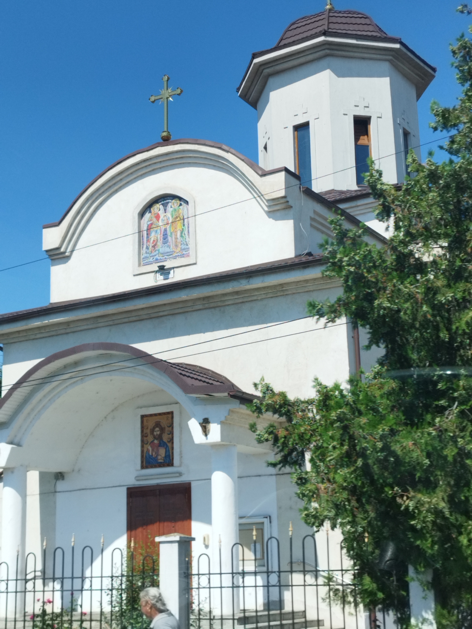 This is a front, ground-level, medium shot of a white church with a brown tiled roof, located in Chitila, Romania. The church has a large archway over the entrance, flanked by white columns. Above the archway, a mosaic depicts figures in colorful robes. A smaller mosaic of Jesus is located above the wooden double doors of the entrance. A tall, octagonal tower with a brown roof is visible on the right side of the church, with tall, narrow windows and decorative cross-shaped indentations. A green metal cross stands on the peak of the church's roof. A black metal fence runs across the front of the property, with a person with gray hair walking behind it. Lush green trees partially obscure the right side of the church. The sky is clear and blue.
