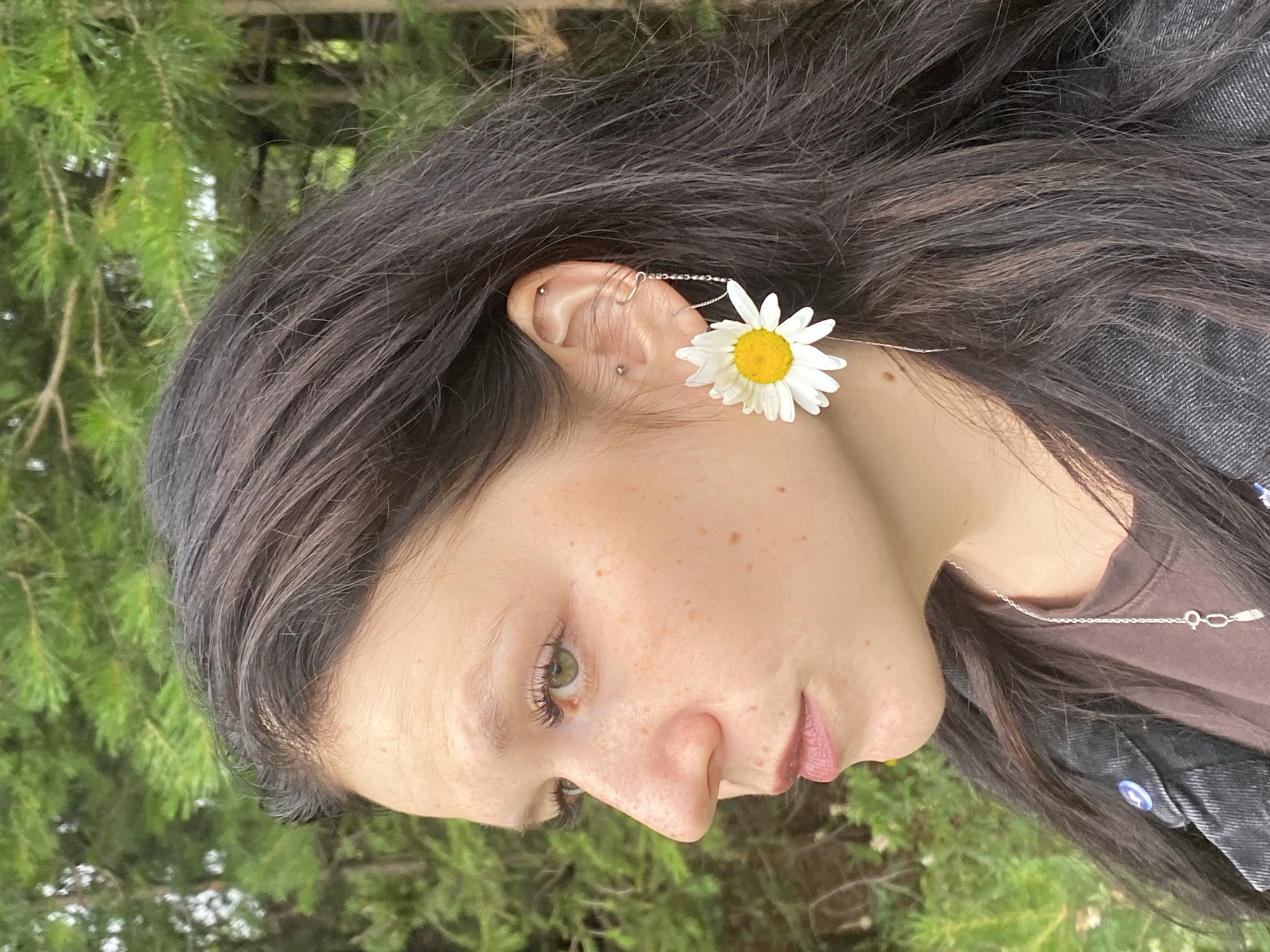 A close-up, sideways portrait of a young person with long, dark hair and freckles across their face. They are wearing a daisy-shaped earring that dangles from their ear and connects with a delicate chain to the top of their ear. The person is also wearing a brown t-shirt and a dark denim jacket with a blue button. They have green eyes and pink lips, and their gaze is directed downwards. The background is blurred and shows greenery, suggesting an outdoor setting. The image appears to have been taken during the daytime, with natural light illuminating the subject. The overall mood is serene and naturalistic. There is no visible text in the image.