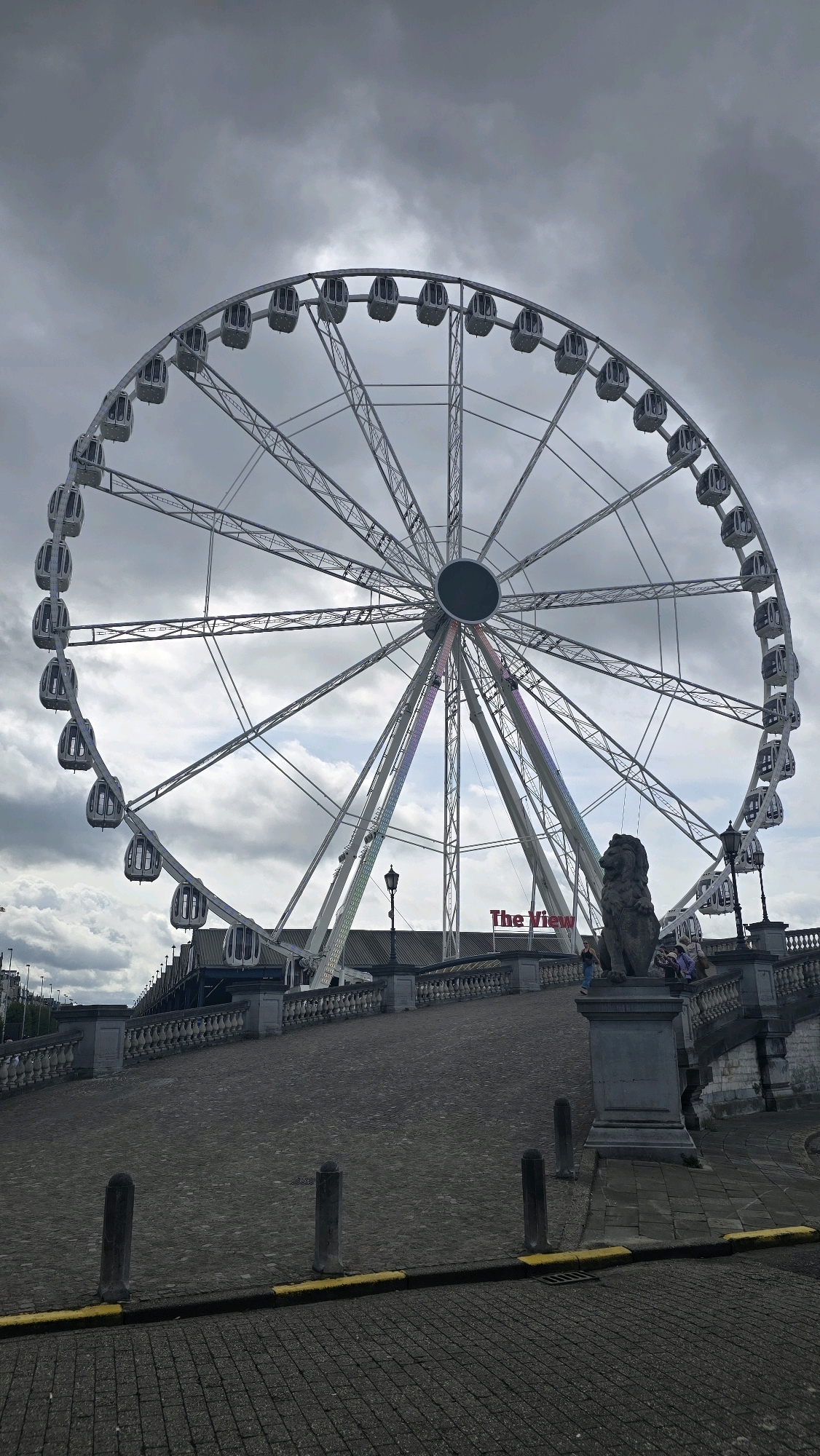 The image captures a large Ferris wheel in Antwerp, Belgium, under a cloudy sky. The Ferris wheel is predominantly white with numerous enclosed gondolas. In the foreground, there is a cobblestone pathway leading up a gentle slope towards a stone balustrade. On the right side of the pathway, there is a stone statue of a lion on a pedestal, and some people are visible walking up the steps and near the statue. In the background, behind the Ferris wheel, the words "The View" are visible in red lettering on a building. Several streetlights are also present along the pathway and near the Ferris wheel. The overall scene suggests an urban environment, likely a tourist attraction or entertainment area.