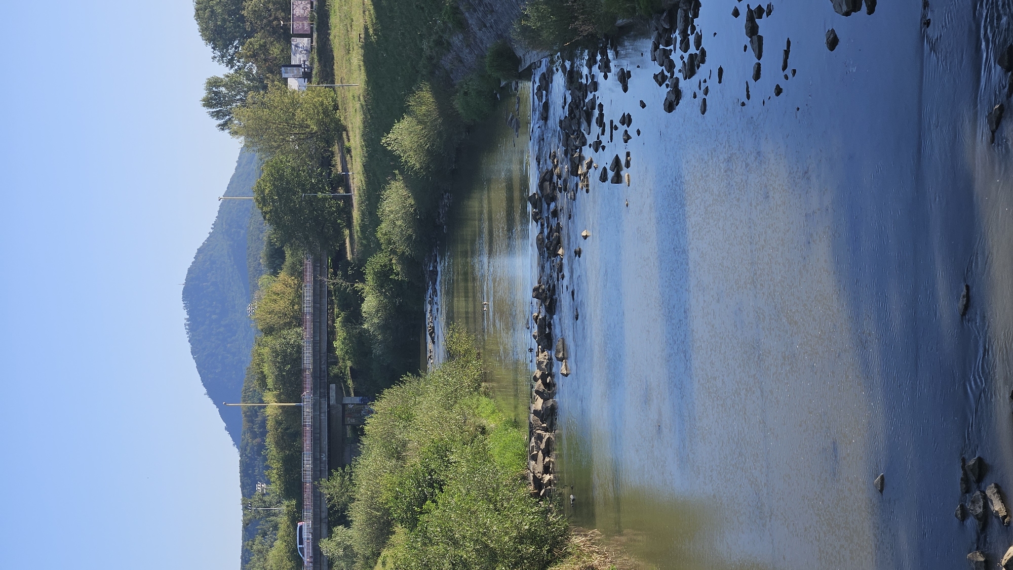 The image depicts a wide, shallow river flowing through a city, with a prominent hill in the background. A concrete bridge spans the river in the middle ground, with vehicles visible on it. The riverbanks are lined with lush green trees and bushes. The water has a greenish-brown hue and is dotted with rocks, some of which form small rapids. The sky is a clear, bright blue, indicating a sunny day. In the distance, the hill, which is characteristic of the Zemplín region, is covered in vegetation and has a small structure on its summit. On the right bank, a grassy slope leads up to a road with some billboards and lampposts. The scene appears to be a natural landscape integrated into an urban environment, possibly a park or a recreational area within the city of Prešov, Slovakia.