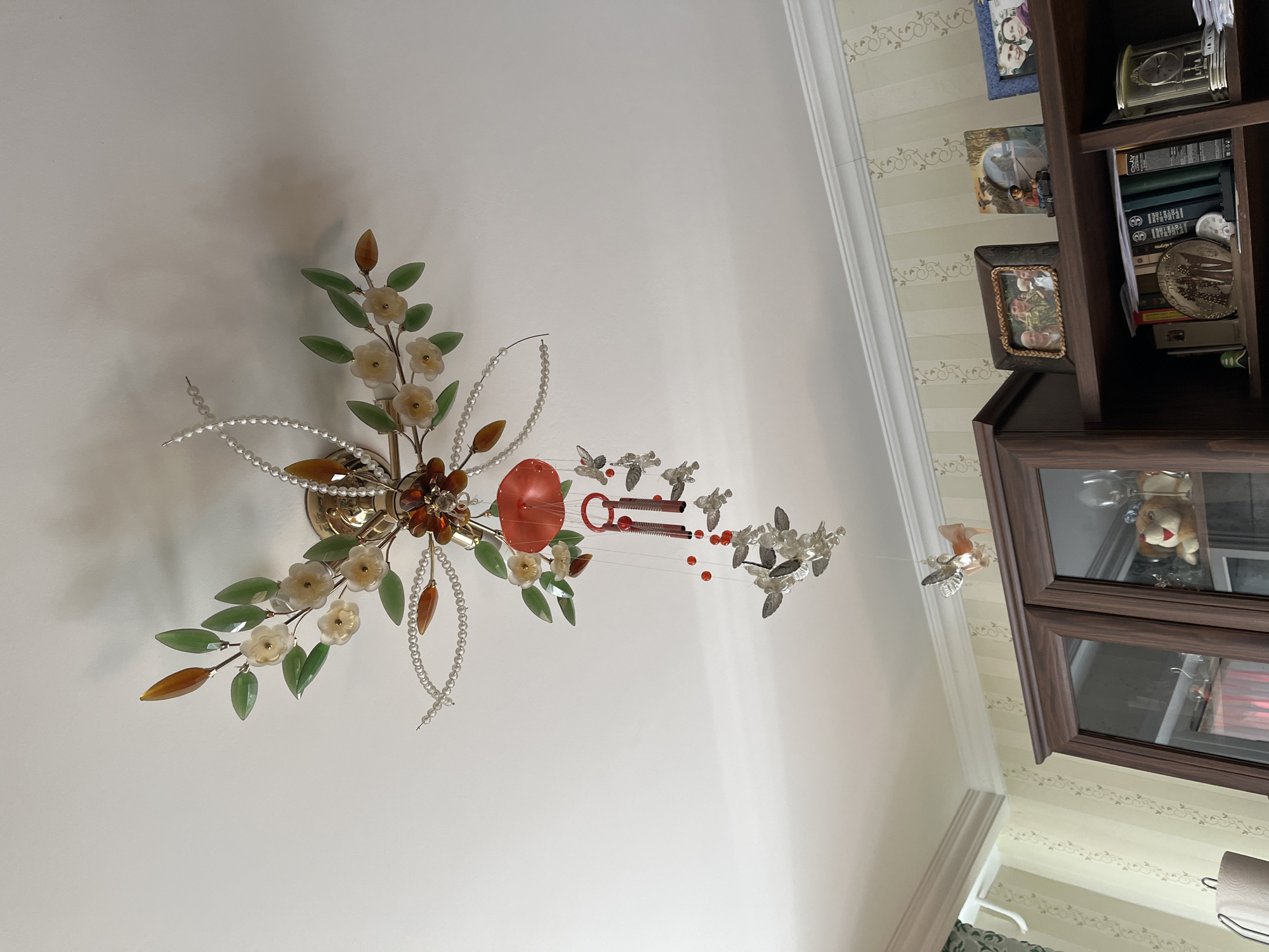 The image depicts a ceiling-mounted decorative light fixture with a floral and pearl design, featuring green glass leaves, small white flowers, and amber-colored accents. Hanging from the fixture is a red, umbrella-shaped ornament, accompanied by several small silver-colored butterflies and a set of chimes. Below the ceiling, a wooden cabinet with glass doors stands against a wall adorned with striped wallpaper. The cabinet's shelves are populated with various items, including framed photographs, books, a vintage clock, and decorative objects like a stuffed animal. The overall scene suggests an indoor residential setting. The location is specified as a city in Minsk, Belarus.