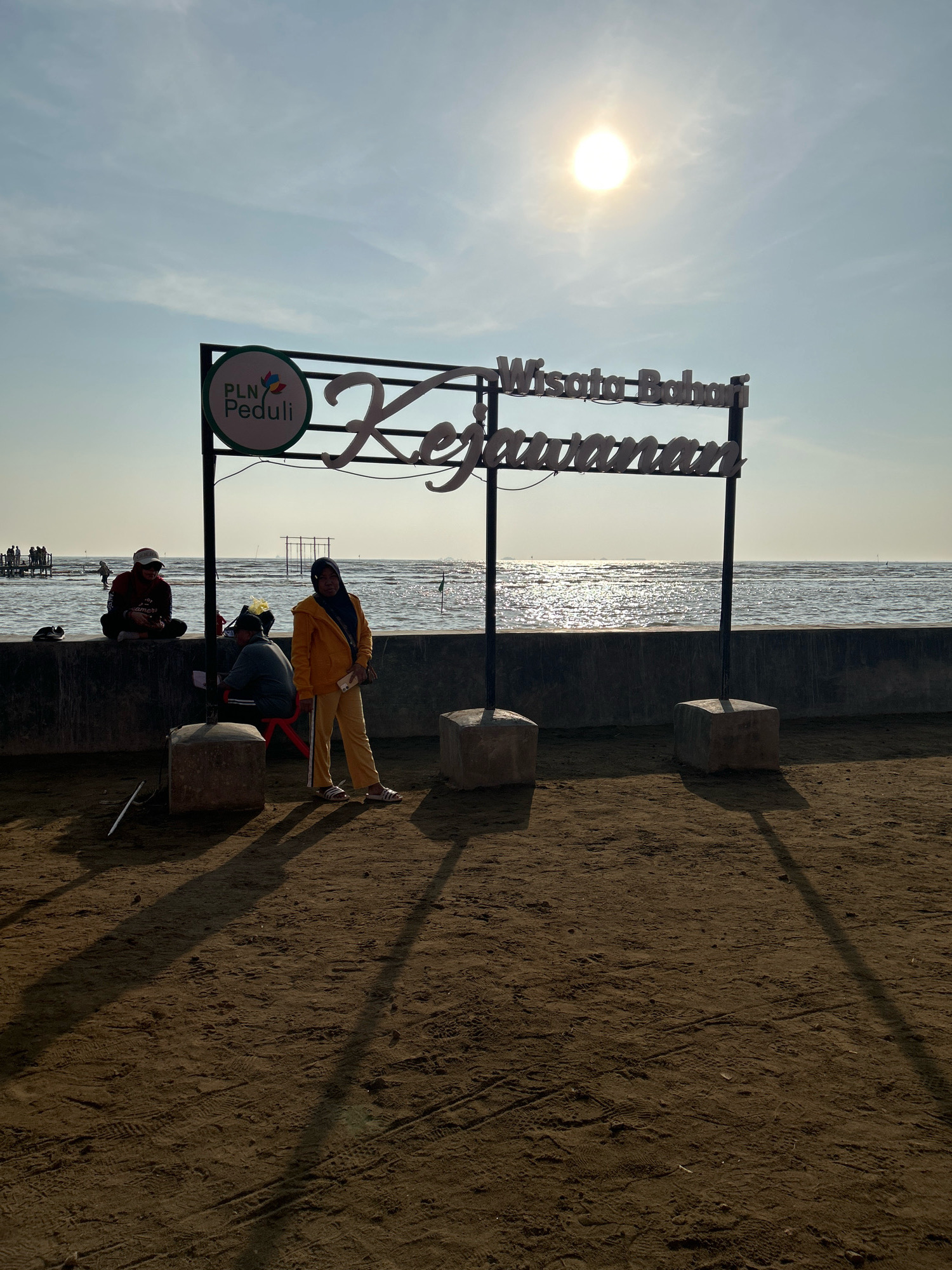 The image depicts the "Wisata Bahari Kejawan" sign in Cirebon, Indonesia, a coastal tourist destination. The sun is visible in the upper right quadrant of the sky, casting a bright glare on the water. The sign is supported by two dark poles and has white cursive lettering. Below the sign, on the sand, a woman in a yellow jacket and pants is standing. To her left, two people are sitting on a concrete wall overlooking the sea, one of whom is wearing a red hijab. The environment is a sandy beach with a concrete barrier separating it from the ocean. In the background, several people are visible near the water's edge, and some structures are present in the sea. The overall scene suggests a sunny day at a beachside attraction.