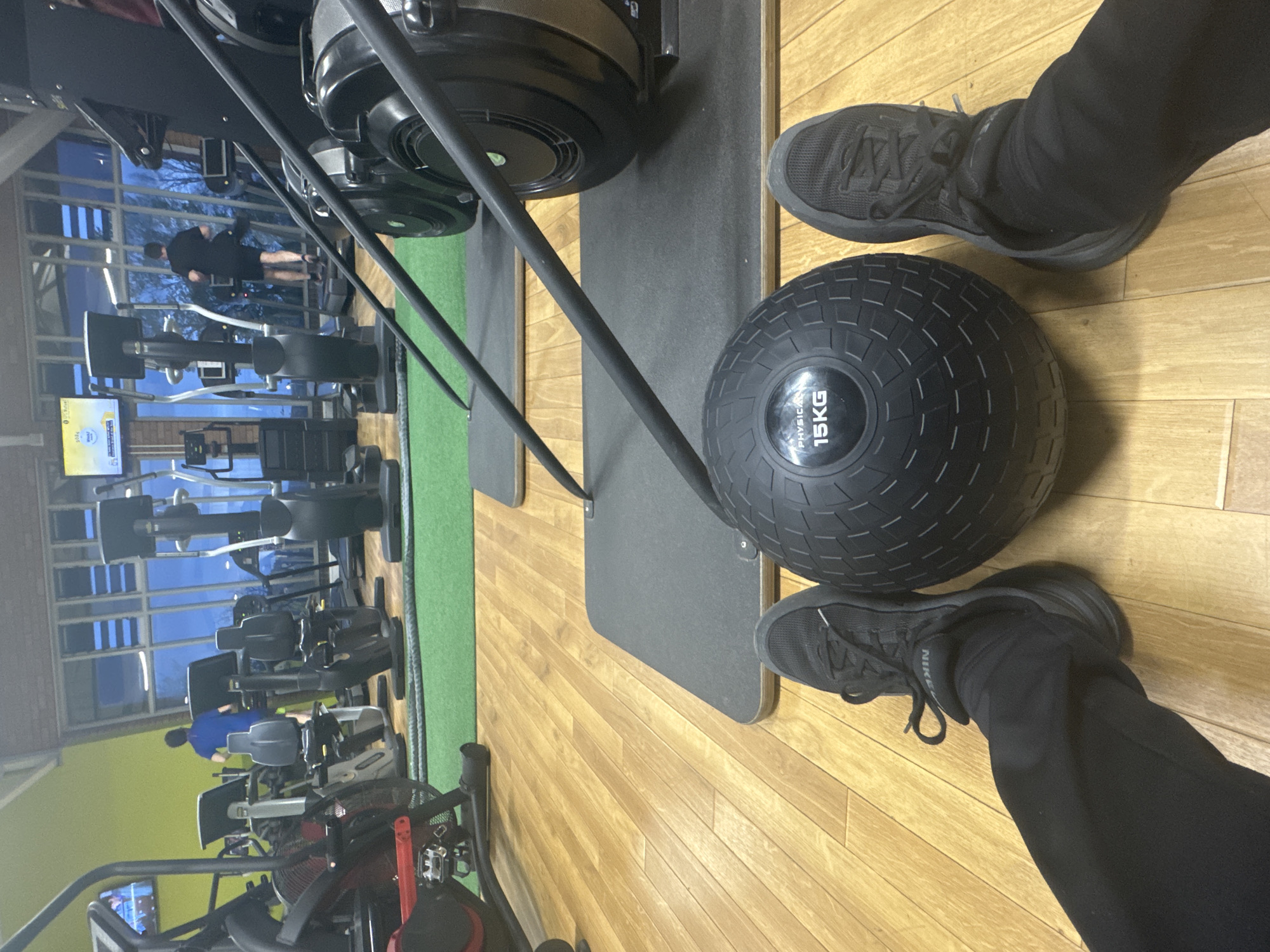 The image shows an indoor view of a fitness center in Welshpool, United Kingdom. The foreground is dominated by a 15kg black medicine ball, flanked by two feet clad in black athletic shoes and black pants. Adjacent to this setup is a rowing machine, with its dark components visible. The floor is constructed of wood panels. In the background, multiple exercise machines are arranged, and individuals are engaged in physical activity. Through a large window, a glimpse of the exterior landscape is visible, including trees and the twilight sky.