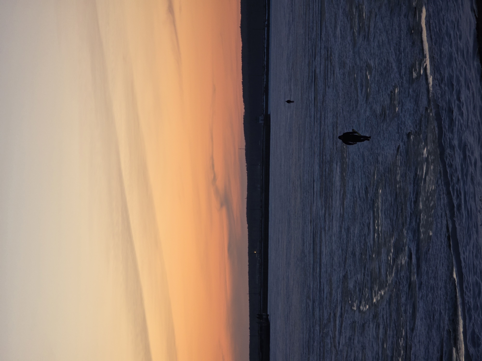 The image captures a sunset view over the water in Chaikovsky, Russia. The sky is a gradient of colors, transitioning from a pale white to a vivid orange. A dark tree line is seen on the horizon, likely on the opposite bank of the body of water. In the water, which is dark blue, two birds are visible. The water surface shows signs of texture, potentially slight ripples or the beginning of ice formation.