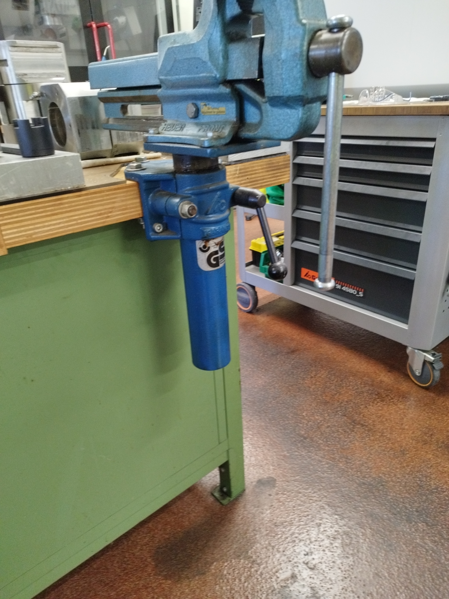 This image depicts a workshop environment in Bremerhaven, Germany. The foreground is dominated by a heavy-duty blue vise attached to the edge of a green workbench. The vise has a long silver handle and a blue cylindrical attachment hanging below it. Behind the workbench, a grey rolling tool cabinet with multiple drawers is visible, partially obscured by the vise and its attachment. The cabinet has various wheels, some with orange accents. The floor is a mottled reddish-brown. In the background, a glass-paneled door or window allows a glimpse into another area with what appears to be some sort of machinery or equipment, including red tubular structures and reflective surfaces. The overall scene suggests a functional, industrial or mechanical workshop setting.