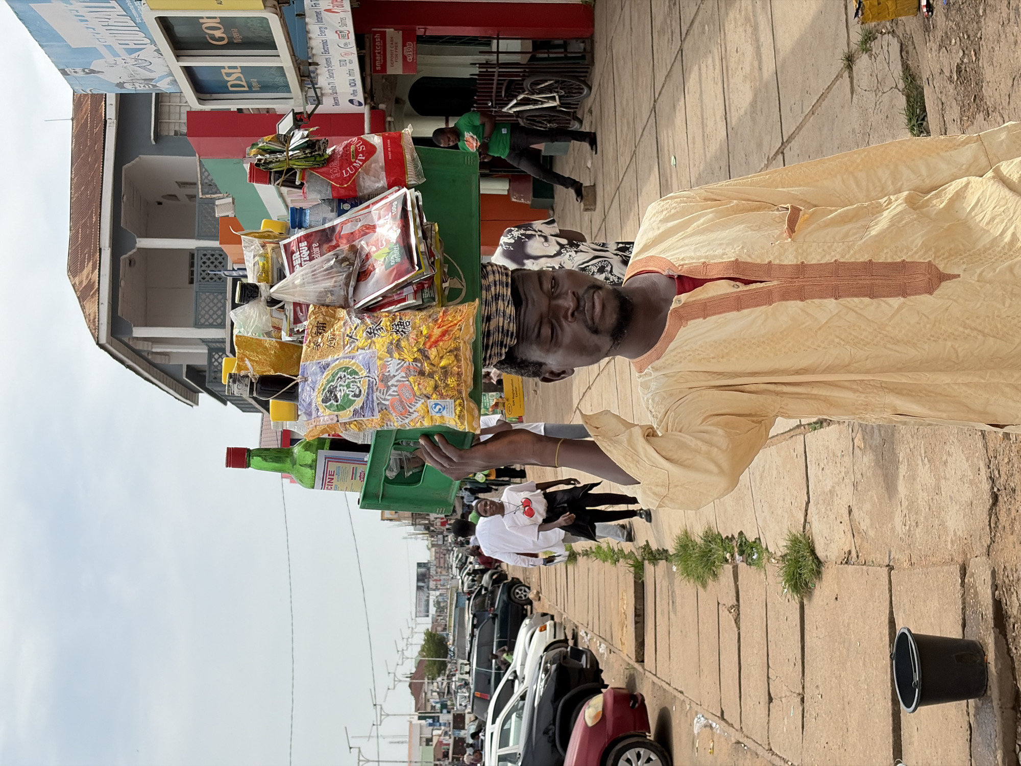 The image depicts a man carrying a load of goods on his head in a street market in Jos, Nigeria. He is wearing a light-colored kaftan with intricate brown embroidery. On his head, he balances a green plastic crate filled with various items, including bottles of liquid, packaged snacks, and other products. The man appears to be walking down a paved sidewalk lined with a grassy median.
In the background, there are buildings, including shops with prominent signage, and parked cars. The street appears to be busy with traffic and pedestrians. One shop has a billboard advertising "DSTV Nigeria" and "GOTV Nigeria". Another sign reads "smartcash DEPOSIT CASH FOR FREE". A man in a green t-shirt and dark trousers is visible looking at his phone, leaning against a fence. A bicycle is also parked nearby. The sky is overcast. The overall scene captures the bustling atmosphere of a market in an urban setting in Nigeria.