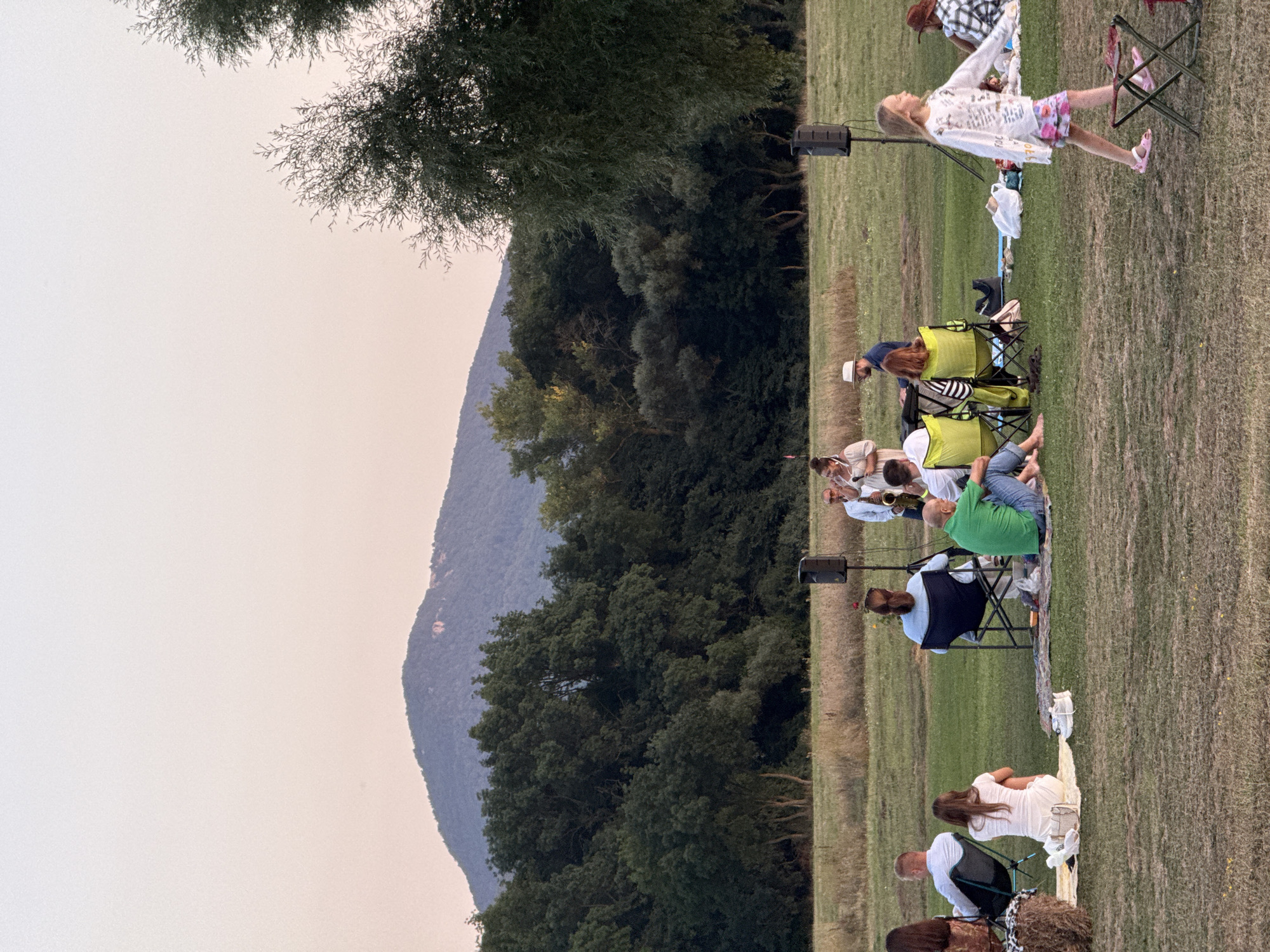 An outdoor concert is taking place on a grassy field with a large mountain and a dense forest in the background. The sky is a pale pink and orange, indicating sunset. A small group of people are seated on chairs and blankets, facing a band that is performing. The band consists of three musicians playing a saxophone, keyboard, and drums. There are two large speakers on stands to the side of the stage. A young girl in a white shirt and pink shorts is walking in the foreground, to the right of the frame. The atmosphere appears relaxed and informal, suggesting a community event. The location is in the Orlinovskiy Okrug, Ukraine.
