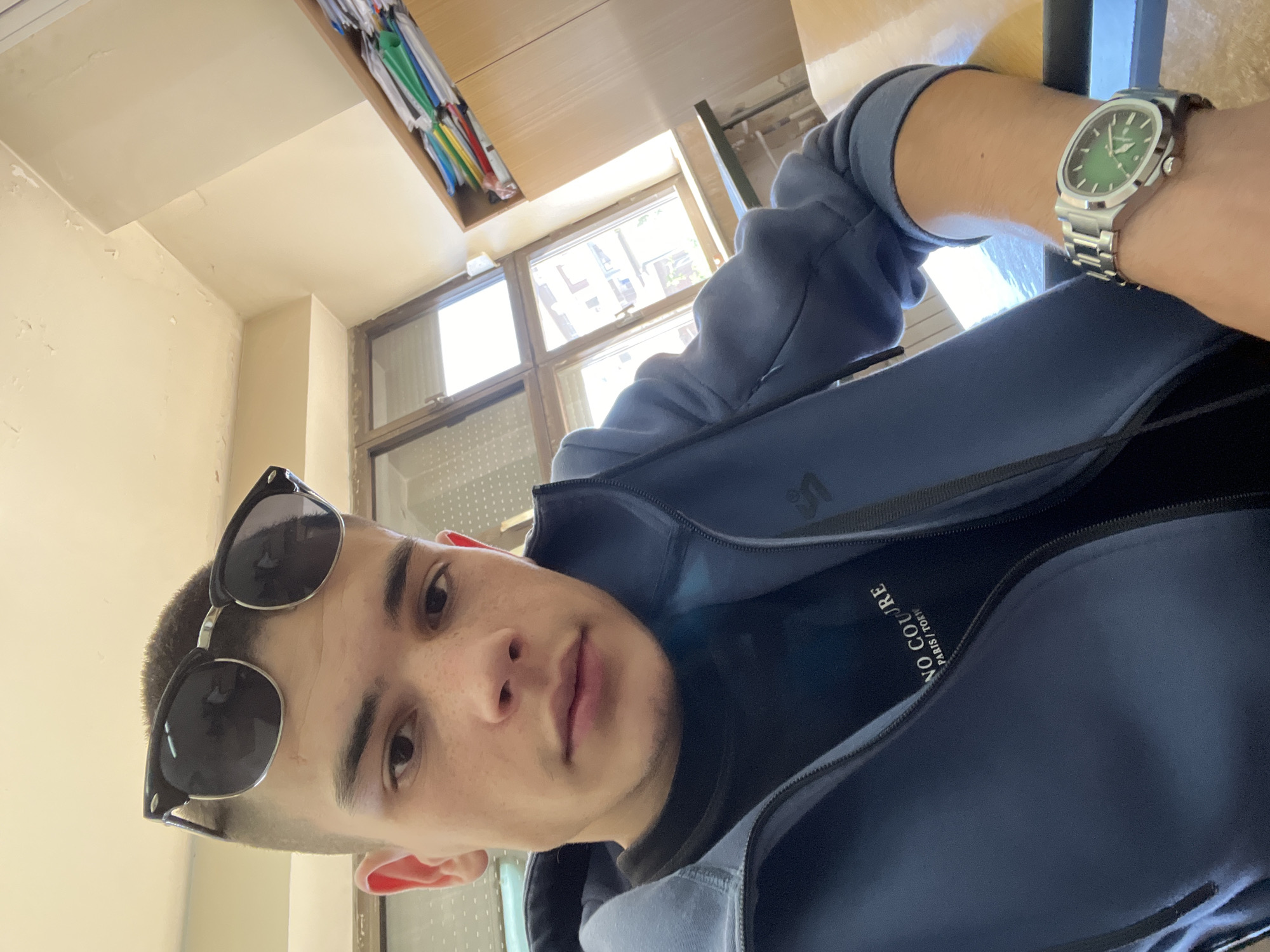 A young man wearing sunglasses and a blue jacket is sitting inside a building with large windows, possibly in Plovdiv, Bulgaria. He has a watch on his wrist. There is a shelf with books or files in the background.