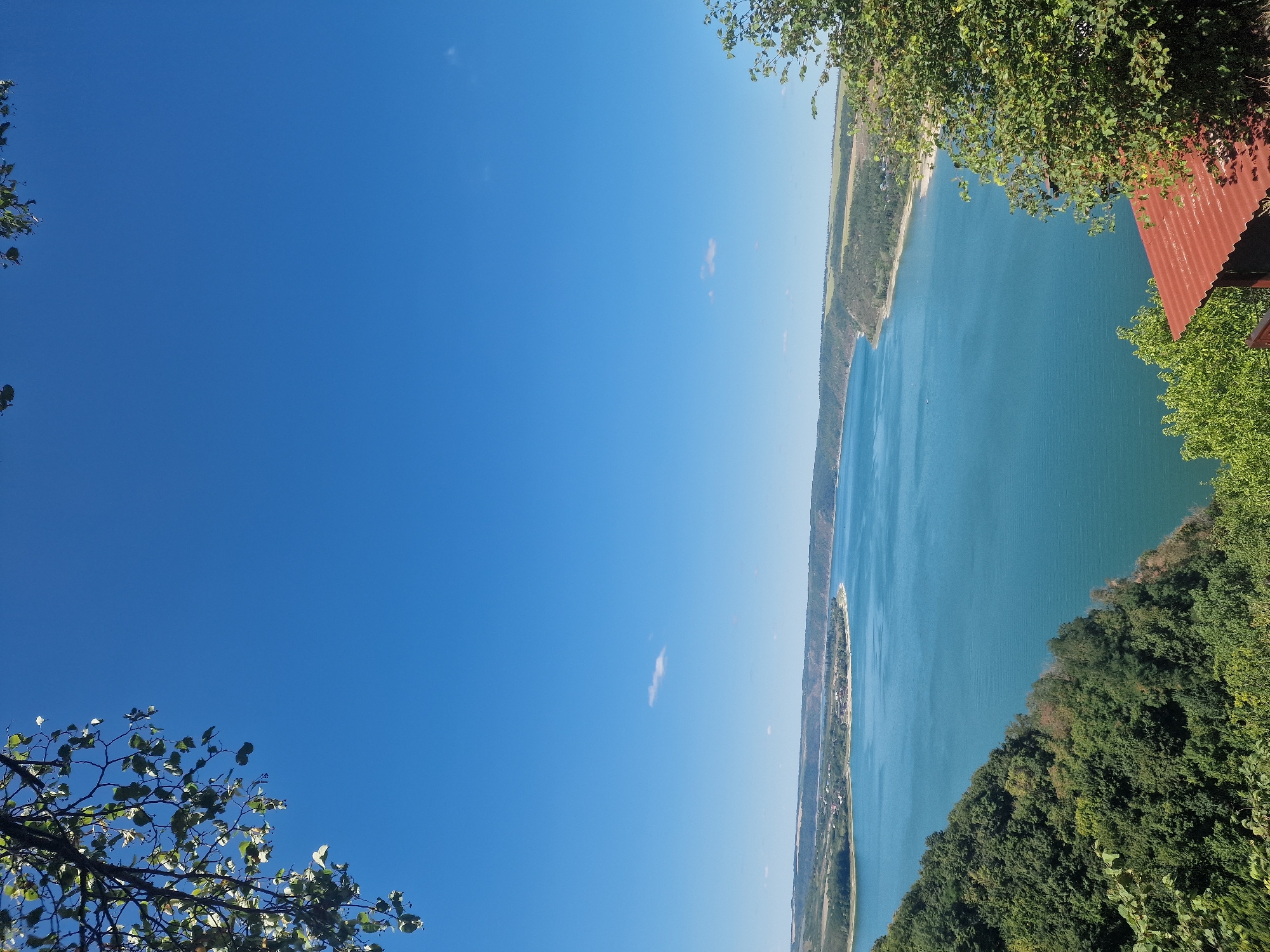 This is a photograph of a wide, tranquil blue river or large lake, seen from an elevated viewpoint. The water stretches into the distance, bordered by rolling hills and a strip of land with some scattered buildings and greenery. The sky above is a clear, bright blue with a few wispy white clouds. In the foreground, the left side of the image is partially obscured by the branches and leaves of a tree, while the right side features dense green foliage and the corner of a rustic, reddish-brown roof of a small structure. The overall scene depicts a natural landscape on a sunny day. There are no people visible in the image. The location is not within the city of Saint-Josse-ten-Noode - Sint-Joost-ten-Node, Belgium, as this is a natural landscape scene far removed from an urban environment.