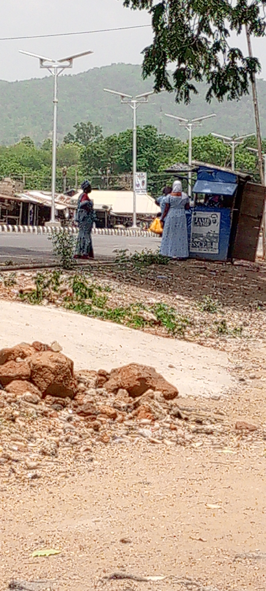 The image captures an outdoor roadside scene in Adankolo, Nigeria, during what appears to be daytime under an overcast or hazy sky.
In the foreground, the ground is unpaved and dusty, covered with loose gravel, small rocks, and sparse green vegetation. A concrete pathway borders this area, leading up to a paved road.
The middle ground features several people and objects along the roadside. A woman in a patterned blue and grey dress and headwrap stands near the road's edge. To her right, another woman, dressed in a light blue or white patterned long dress and white head covering, is positioned beside a small, blue kiosk or vendor stall. She seems to be interacting with the stall, which displays the word "LAMP" and a graphical illustration, possibly a product, on its side. Another individual is partially visible behind her, holding what looks like a yellow bag.
The paved road has a distinctive black and white striped curb. Across the road, various makeshift structures or small buildings are visible, suggesting a commercial or market district. Modern streetlights, identifiable by their prominent solar panels, are spaced along the roadside. The background showcases large, verdant hills or mountains, indicative of the natural landscape surrounding the area. No specific activity or event beyond everyday roadside interactions is clearly discernible.