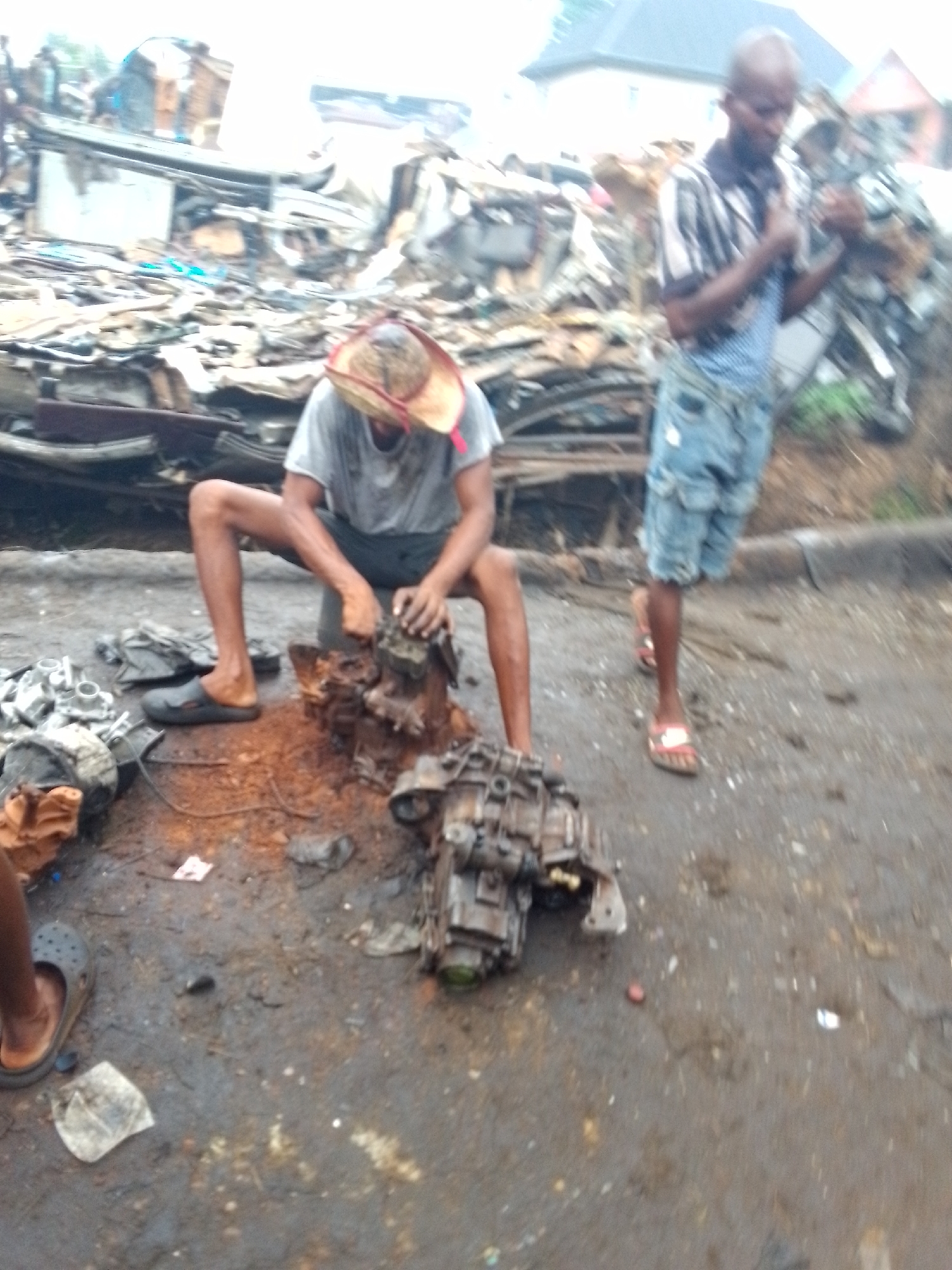 The image depicts a scene in an outdoor, informal setting, likely a scrap or salvage yard, in Onitsha, Nigeria. Two men are the primary subjects, with a third person partially visible.
In the foreground, a man wearing a light-colored t-shirt and a straw hat with a red band is squatting low to the ground. He is intensely focused on disassembling or working on what appear to be heavily soiled and corroded mechanical components, possibly from a vehicle engine or transmission. Several other metallic parts are scattered around him. One of his feet is bare, while the other wears a dark slip-on shoe.
To the right, a second man stands, facing slightly away from the camera. He wears a striped short-sleeved shirt, blue denim shorts, and red flip-flops. He appears to be holding and looking down at a small object in his hands, possibly a mobile phone.
The ground is uneven, muddy, and wet, suggesting recent rainfall or damp conditions. The overall background is dominated by large, haphazard piles of scrap metal, wood, and various debris, forming a cluttered, industrial landscape. In the far background, glimpses of buildings, including one with a dark roof, can be seen through the haze and clutter. The lighting is diffused, indicating an overcast day. No visible text is discernible in the image. The scene conveys an atmosphere of labor and resourcefulness within a challenging environment.