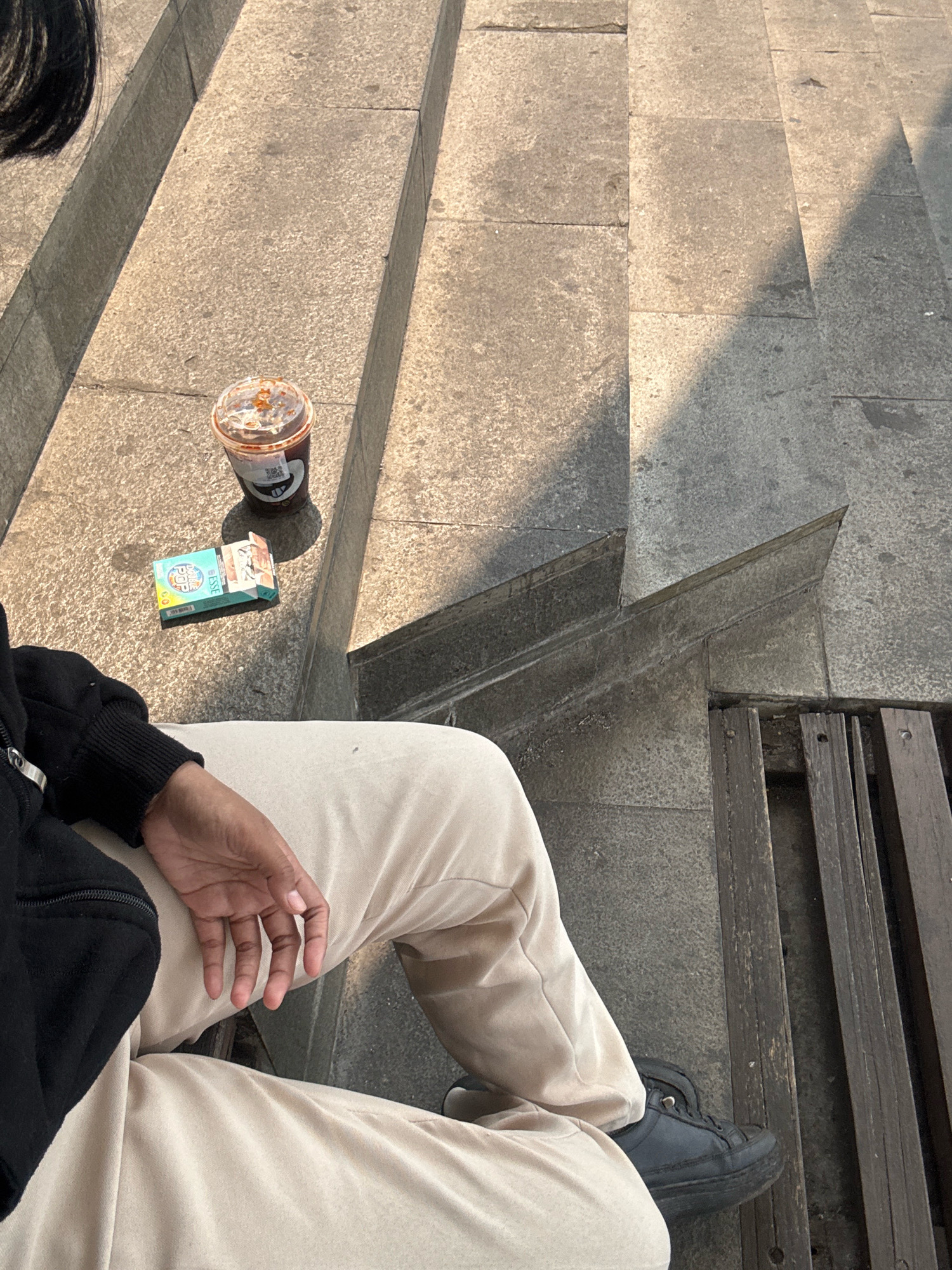 A person is sitting on concrete stairs outdoors, with a pack of ESSE cigarettes and a cup of iced coffee placed on the step beside them. The person is wearing a black jacket and light-colored pants. A wooden bench is visible to the right of the stairs. The scene suggests a moment of relaxation in an urban environment, possibly in North Jakarta, Indonesia, given the presence of ESSE cigarettes which are popular in Indonesia. The lighting indicates it is daytime, with sunlight casting shadows on the stairs.