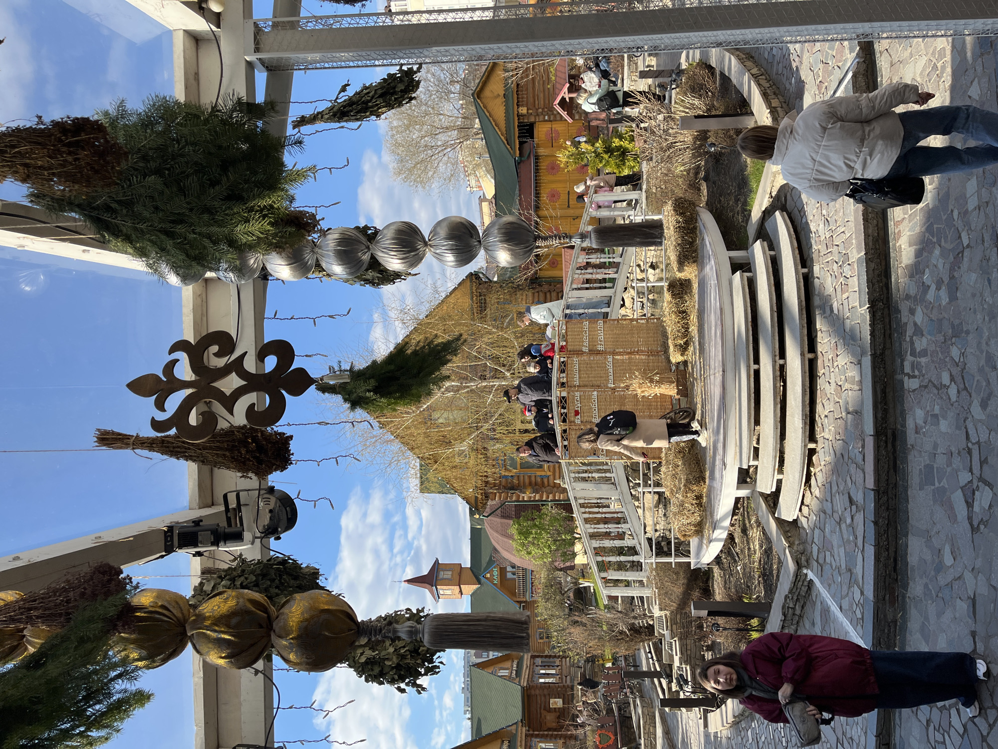 This image captures an outdoor scene in Kazan, Russia. The environment is a street with a mosaic cobblestone pavement. The architecture is dominated by wooden structures, including houses and buildings, some with a Russian traditional style. People are visible in the background, some on a raised platform, suggesting a cafe or gathering place. Decorative elements are suspended from an overhead structure, including ornaments, greenery, and lights. A woman stands in the foreground. There are also benches made of white stone or concrete. The sky is partly cloudy with blue sky peeking through. Text is visible on a side panel, written in Russian, with the word “Cafe”.