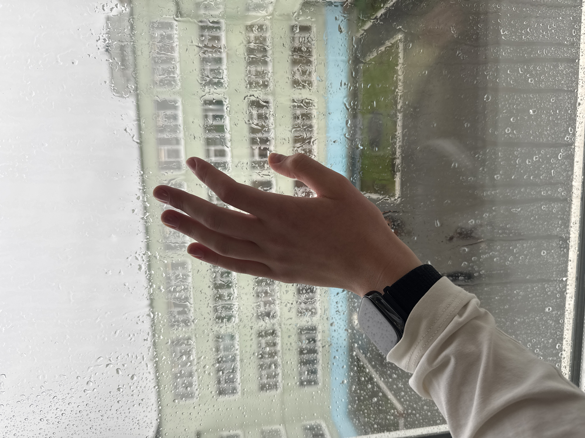 The image presents a close-up indoor perspective, looking out through a window heavily covered in raindrops and streaks. A human hand, likely female, is prominent in the foreground, reaching towards the wet glass. The fingers are slightly spread, and the hand appears to be gently touching or gesturing towards the rain-streaked pane. The person's forearm, clad in a white long-sleeved shirt, is visible extending from the right side of the frame. A dark smartwatch with a black band is worn on the wrist.
Through the rain-distorted window, the blurred exterior of a multi-story building is discernible in the background. This building features a light green or pale grey facade with multiple rows of rectangular windows. To the right of this primary structure, a bluish-green element, possibly another section of the building or an adjacent structure, is faintly visible.
The scene depicts a rainy daytime, characterized by the abundant water droplets and runoff on the window, indicating ongoing or recent precipitation. The background is significantly blurred due to both the rain and the depth of field, suggesting an urban or residential setting in Chaikovsky, Russia. The image conveys a quiet moment of observation or connection with the outside weather from an interior space. There is no visible text.