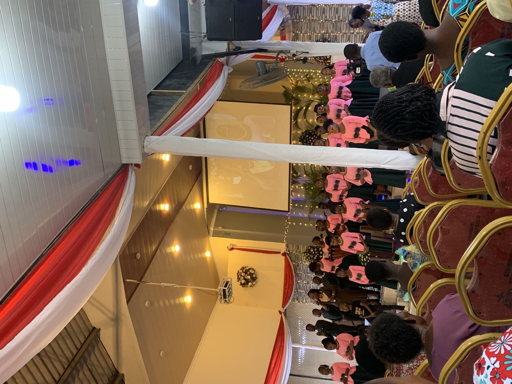A gathering occurs indoors, possibly in Dar es-Salaam, Tanzania, as inferred from the general context. A group is dressed in pink attire; their arrangement suggests a choir or ensemble. Patrons are seated in chairs, positioned to face the stage or performance area. Two projector screens are visible, indicating the presence of AV technology. The venue is adorned with white and red draping, which runs across the ceiling. Lighting is provided by numerous overhead bulbs, and additional decorative lights are interspersed around the stage.