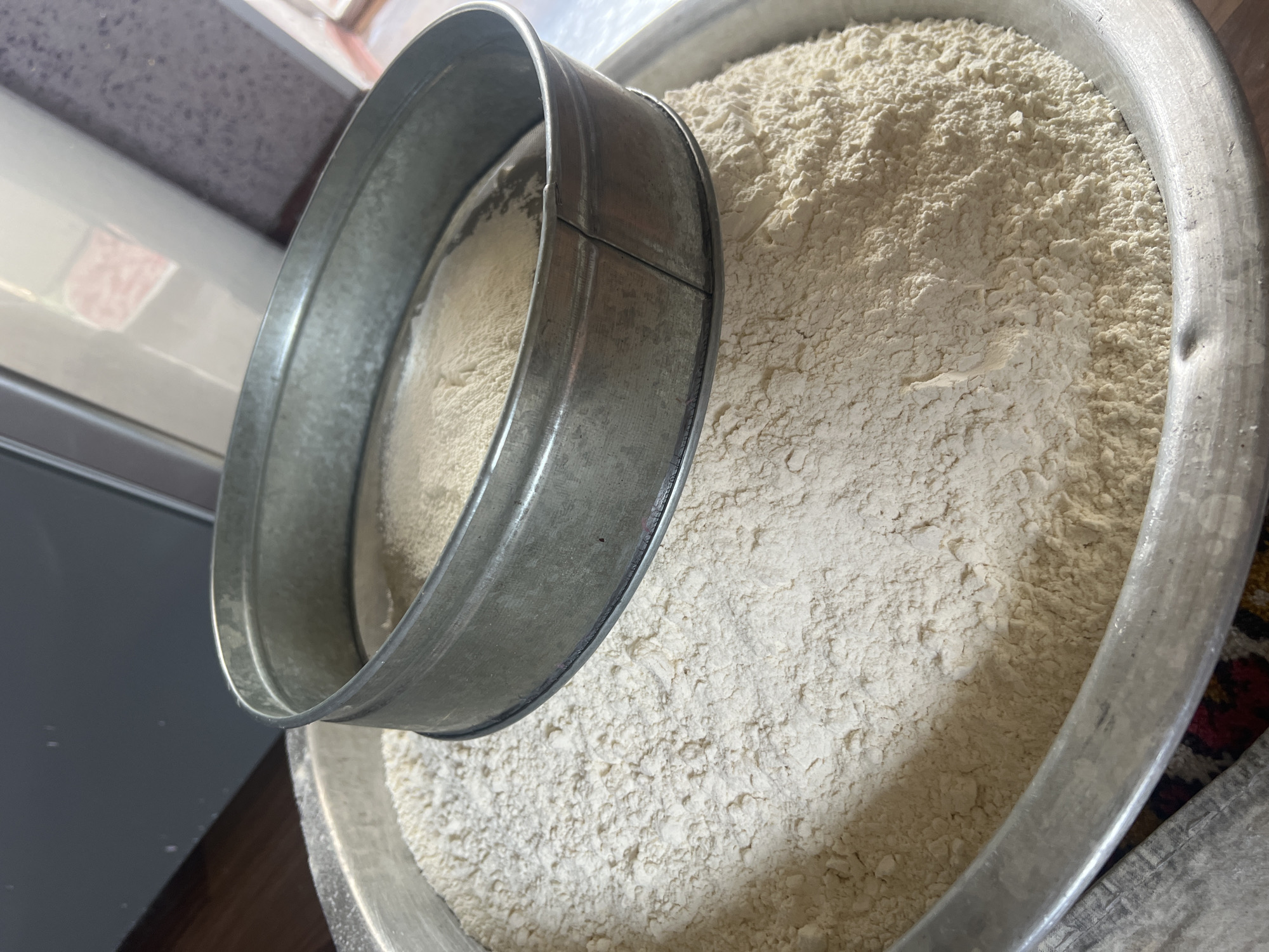In Kara-Dıykan, Kyrgyzstan, a metal sieve rests atop a large metal bowl filled with white flour. The sieve, partially obscuring the flour within the bowl, also has some flour resting on its mesh. The bowl itself appears to be filled to a significant level with the fine powder. The scene is likely indoors, possibly in a kitchen or food preparation area, given the presence of kitchenware.