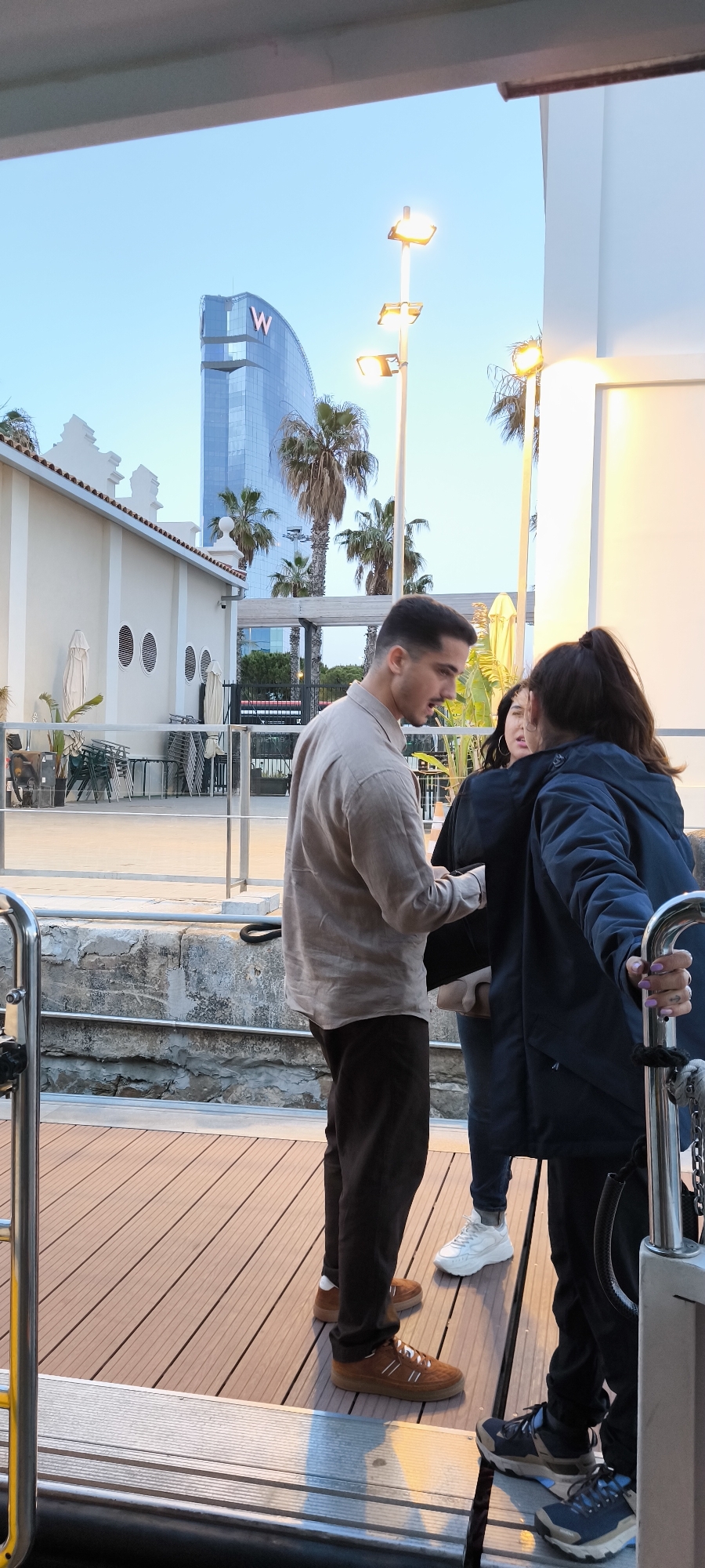 Three people stand on a wooden platform outdoors near a waterfront in Barcelona, Spain. A man in a tan shirt and brown pants faces right, engaging in conversation with a woman in a blue jacket who faces left, holding onto a silver railing. Another woman stands behind them, partially obscured. In the background, there is a building, palm trees, and a structure with a large "W" logo, consistent with the W Barcelona hotel. To the left, there is a building with round windows and stacked chairs, likely a cafe or restaurant. The time appears to be late afternoon or early evening, with soft, muted lighting.