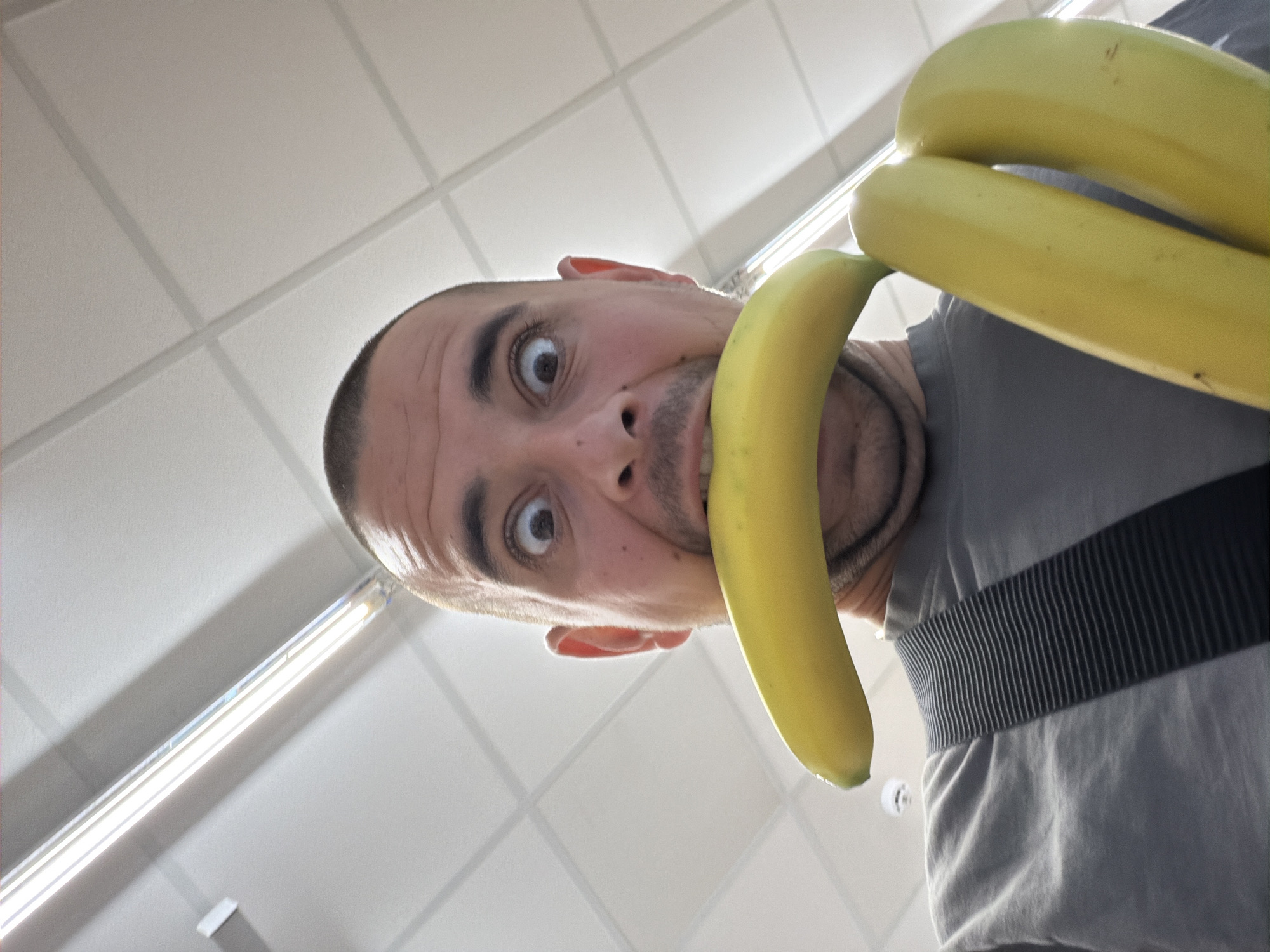 A man is taking a selfie in what appears to be a grocery store, possibly in Burgas, Bulgaria, given the lighting and ceiling tiles. He is holding two bananas in his right hand and has one banana in his mouth, which is open wide with his eyes bulging. He is wearing a grey t-shirt with a black and grey striped strap over his shoulder. The background shows a white tiled ceiling with fluorescent lighting fixtures.