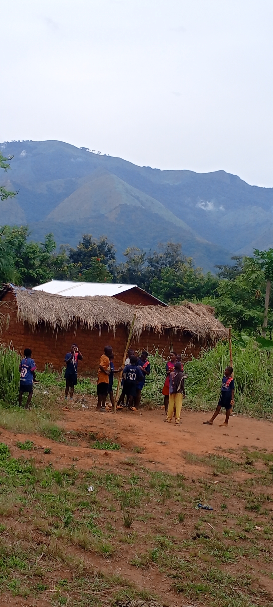 The image depicts a vibrant rural scene in Ngalula, Democratic Republic of the Congo, with a group of children gathered near a traditional building, set against a backdrop of lush mountains.
In the foreground, a reddish-brown dirt path or clearing is visible, with green grass and weeds sparsely growing along its edges.
Approximately eight to ten children, predominantly boys, are the central focus, clustered in the midground. They are dressed casually, with several wearing popular football jerseys. Visible text on these jerseys includes "Neymar Jr 10" on a blue shirt worn by a child on the far left, and "Messi 30" on another dark blue shirt worn by a child towards the center. Part of "Qatar Airways" is also visible on a jersey. The children are gathered around two tall, thin wooden poles, which are vertically staked into the ground, suggesting a makeshift play area or a game. Their postures indicate interaction and play.
Behind the children stands a rustic, rectangular building with brick or mud-brick walls and a prominent thatched roof made of dried straw or grass. To the left and slightly behind this building, a portion of another structure with a white metal roof is visible.
The landscape surrounding the buildings and children is rich with dense, green vegetation, including tall grasses, bushes, and various trees. In the background, majestic mountains rise, their peaks partially obscured by low clouds or mist under an overcast sky, creating a soft, diffused light. The overall impression is of daytime under cloudy or hazy conditions. The scene portrays a moment of childhood activity within a natural and undeveloped setting.