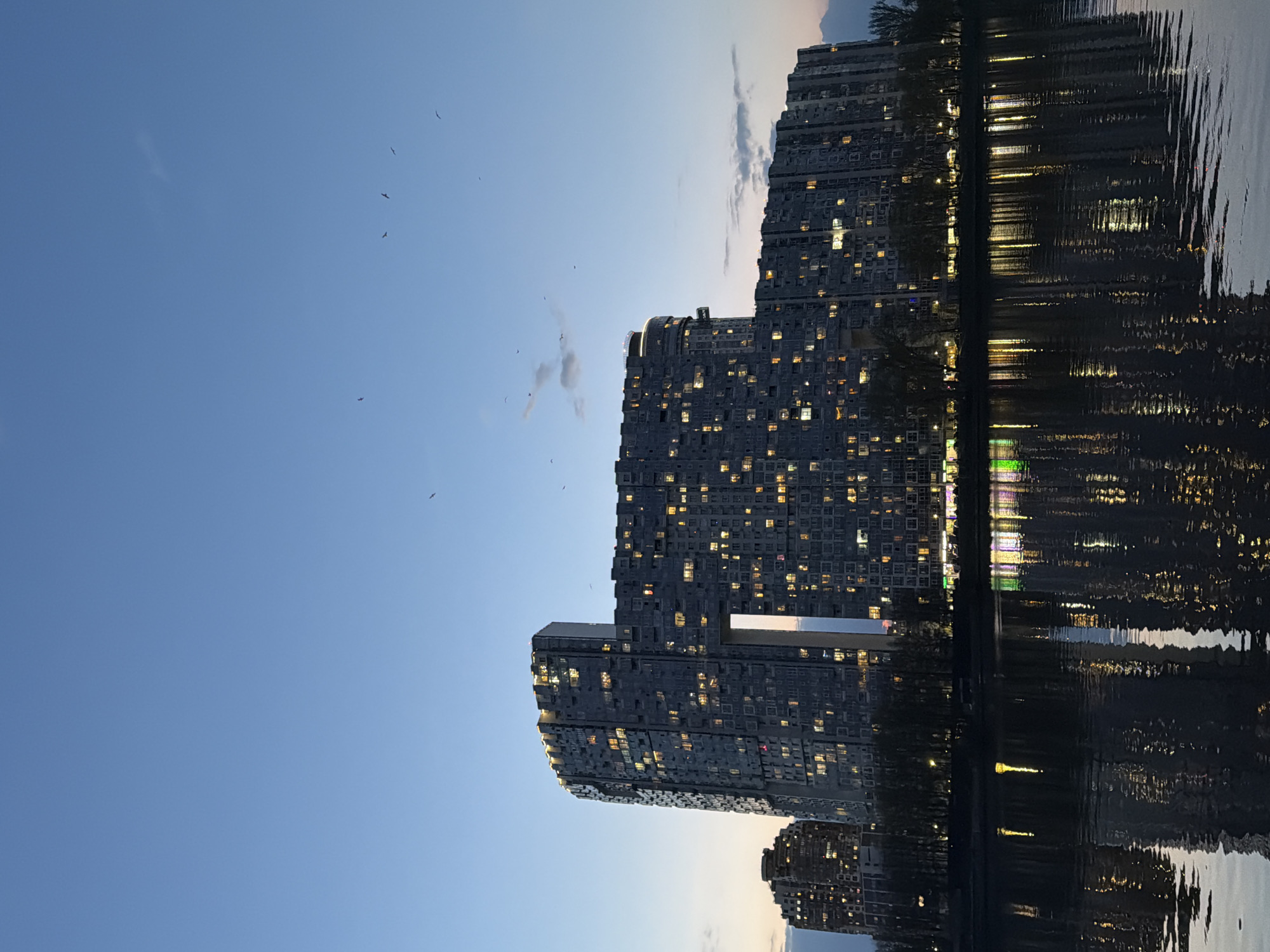 The image depicts a cluster of tall residential buildings reflected in a body of water, likely a lake or river. The buildings appear to be high-rise apartments with numerous windows, many of which are illuminated, suggesting it is evening or dusk. There are birds visible in the sky. The reflection in the water mimics the building lights and architectural details. The scene is possibly in Khimki, Russia, based on architectural features that match the area. The environment is urban, near a waterfront.