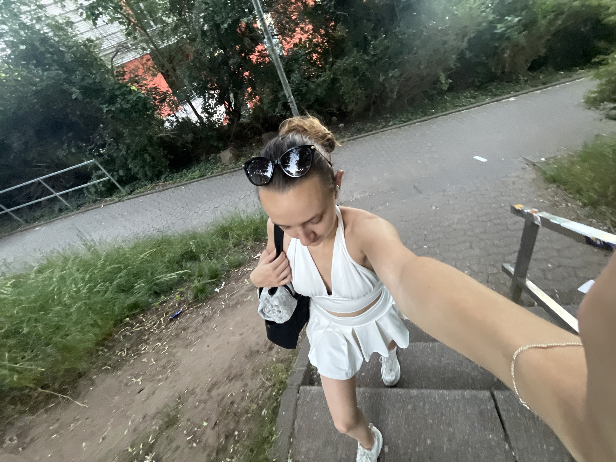 A woman is taking a selfie while descending outdoor stairs in Saarbrücken, Germany. She is wearing a white halter top and skirt, white sneakers, and sunglasses on top of her head. She carries a black bag on her shoulder. The stairs are located in a paved area with greenery and trees visible in the background. A railing is partially visible on the right side of the frame. The light suggests it is daytime.